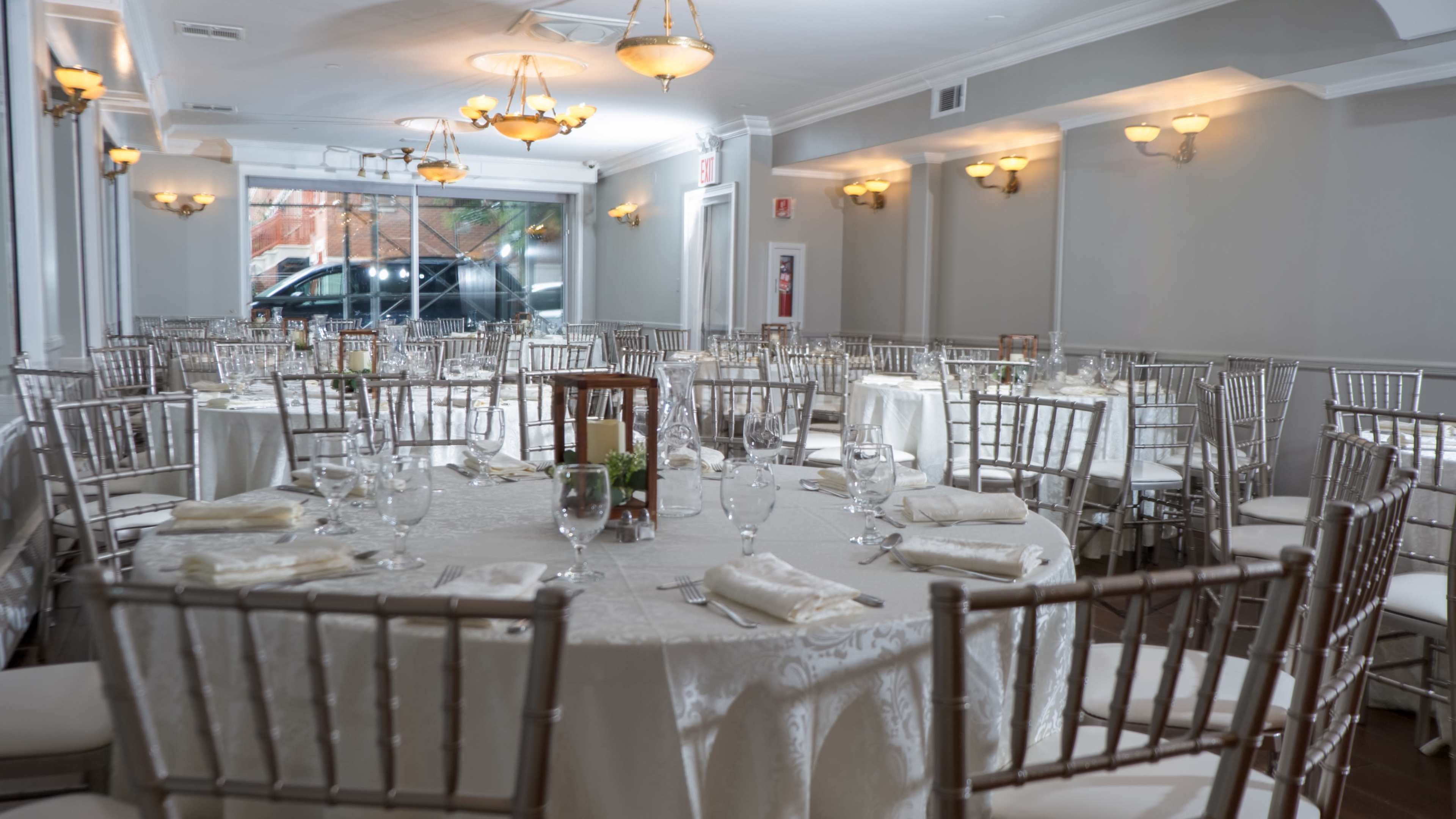 The image shows a banquet room arranged with multiple round tables, each covered with white tablecloths and set with glassware and napkins, illuminated by soft overhead lighting.
