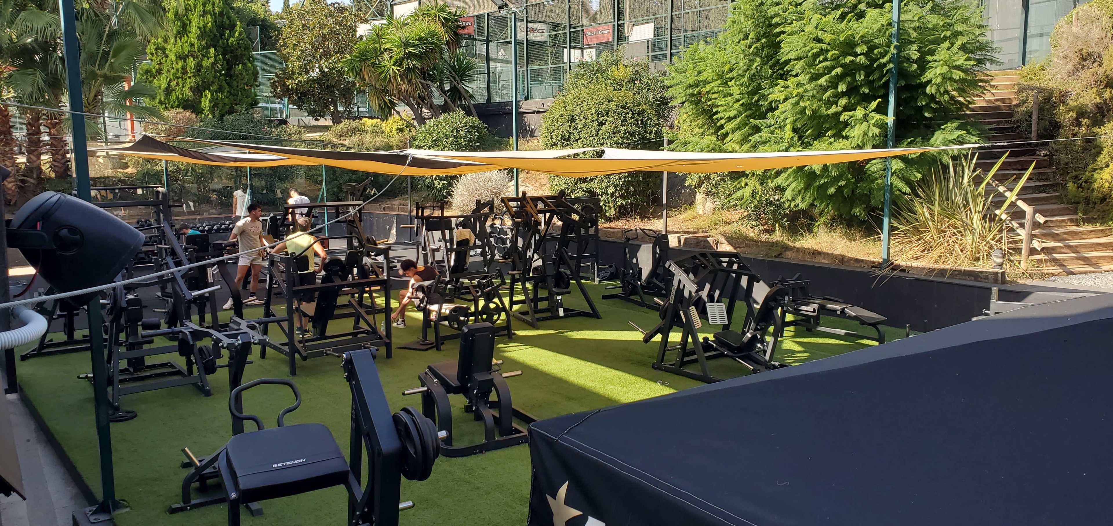 A partially shaded outdoor gym is equipped with various workout machines and weights, surrounded by greenery and a staircase in the background.