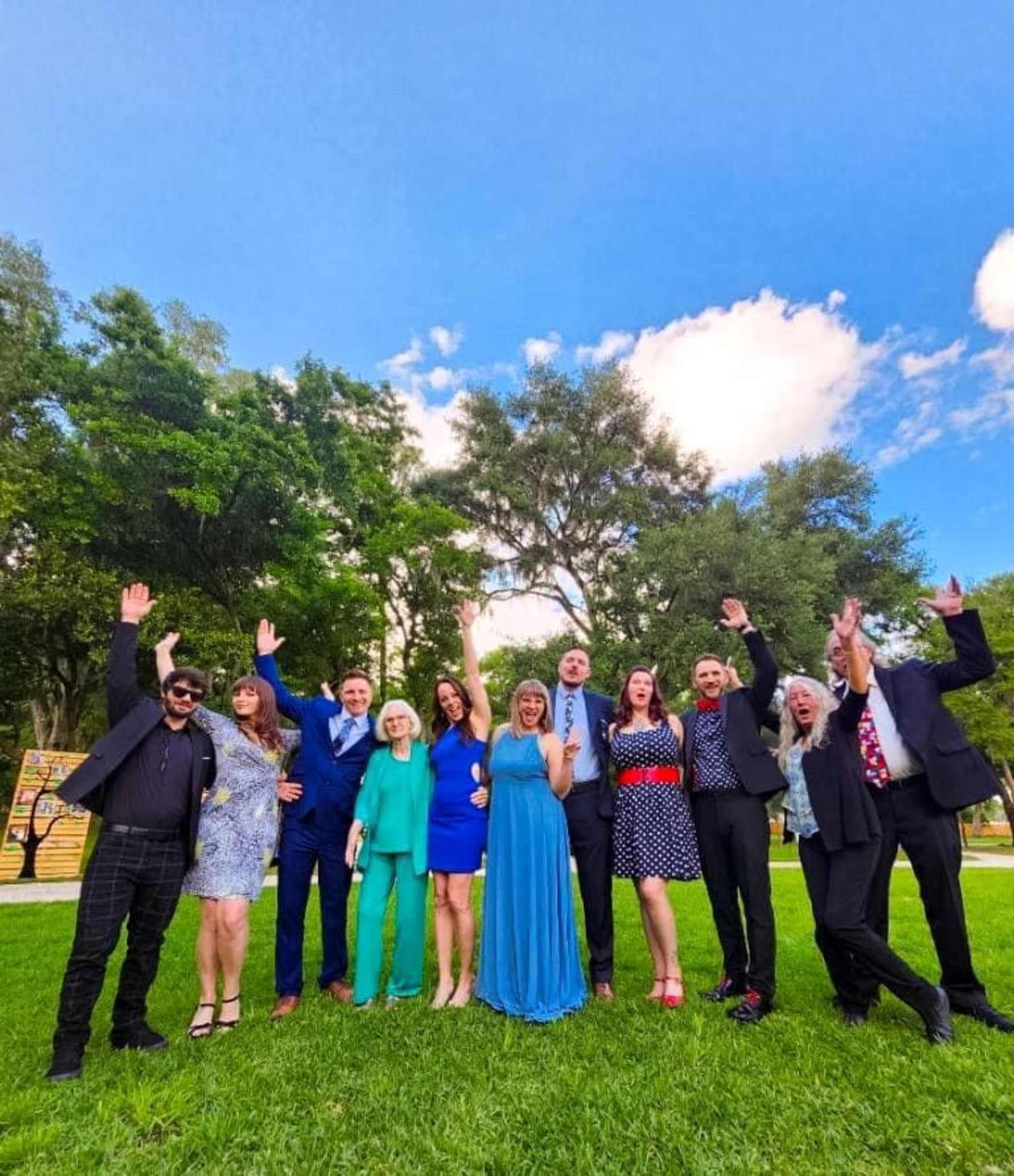 A group of eleven people stands in a park with their arms raised, smiling against a backdrop of greenery and a blue sky.