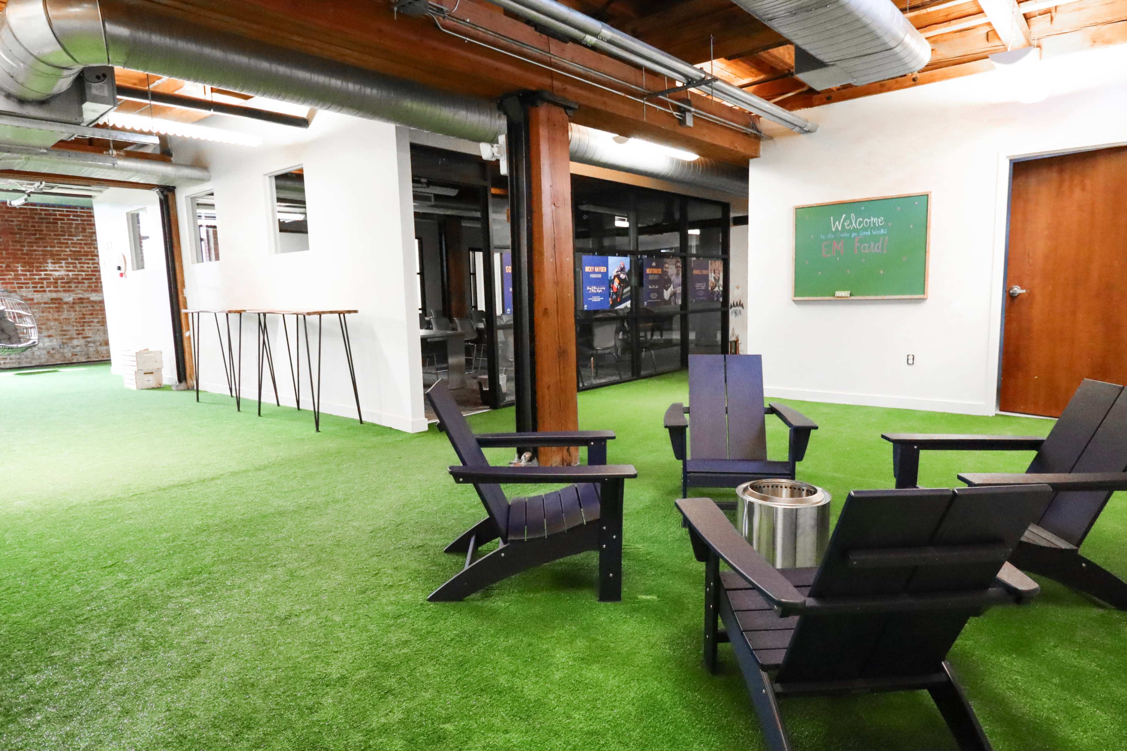 The image depicts a modern office space featuring artificial grass flooring, wooden beams, and a seating area with black Adirondack chairs surrounding a fire pit.