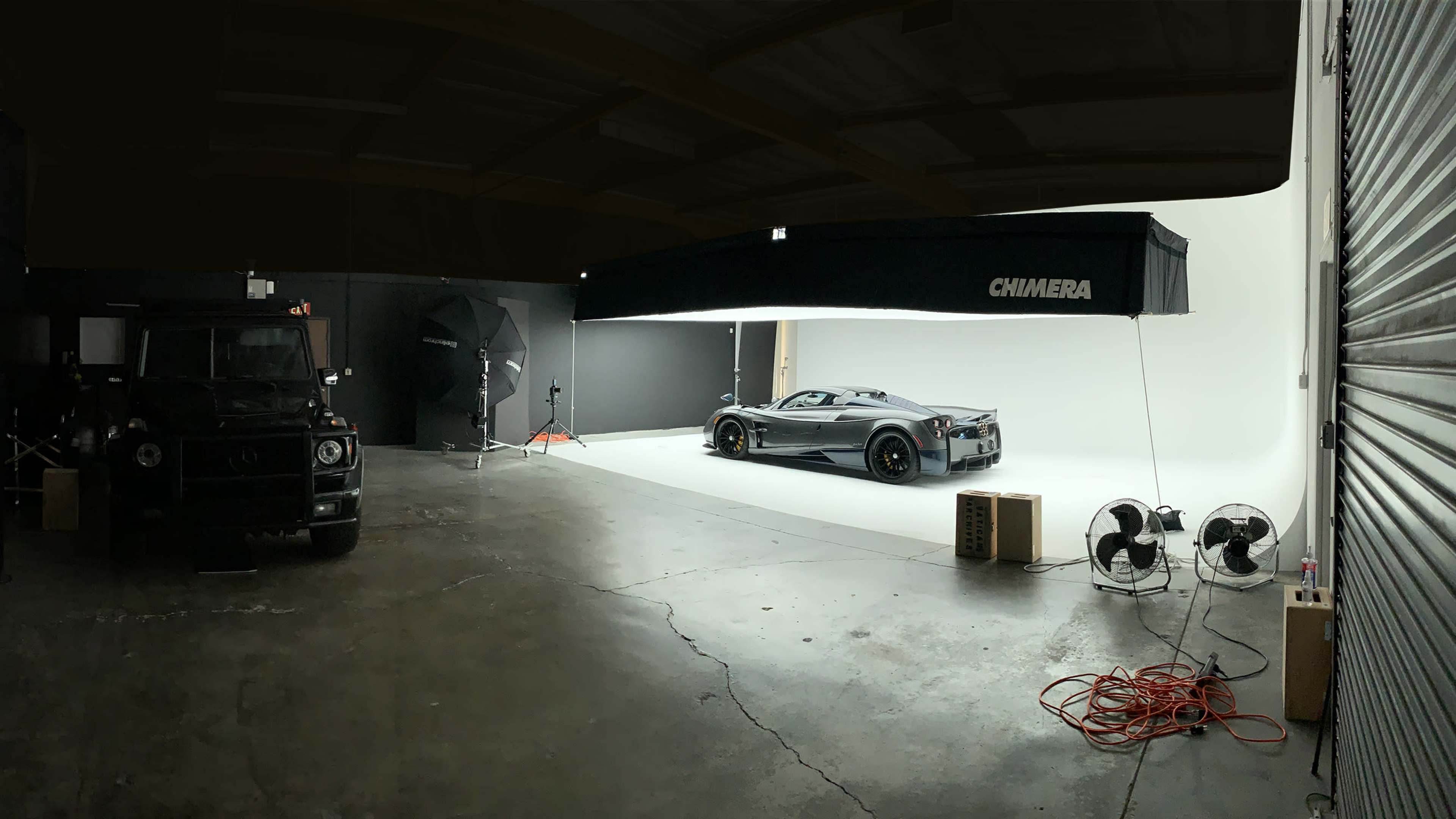 A sleek sports car is positioned on a photography stage with bright white backdrops and studio equipment, while a dark SUV is parked nearby.