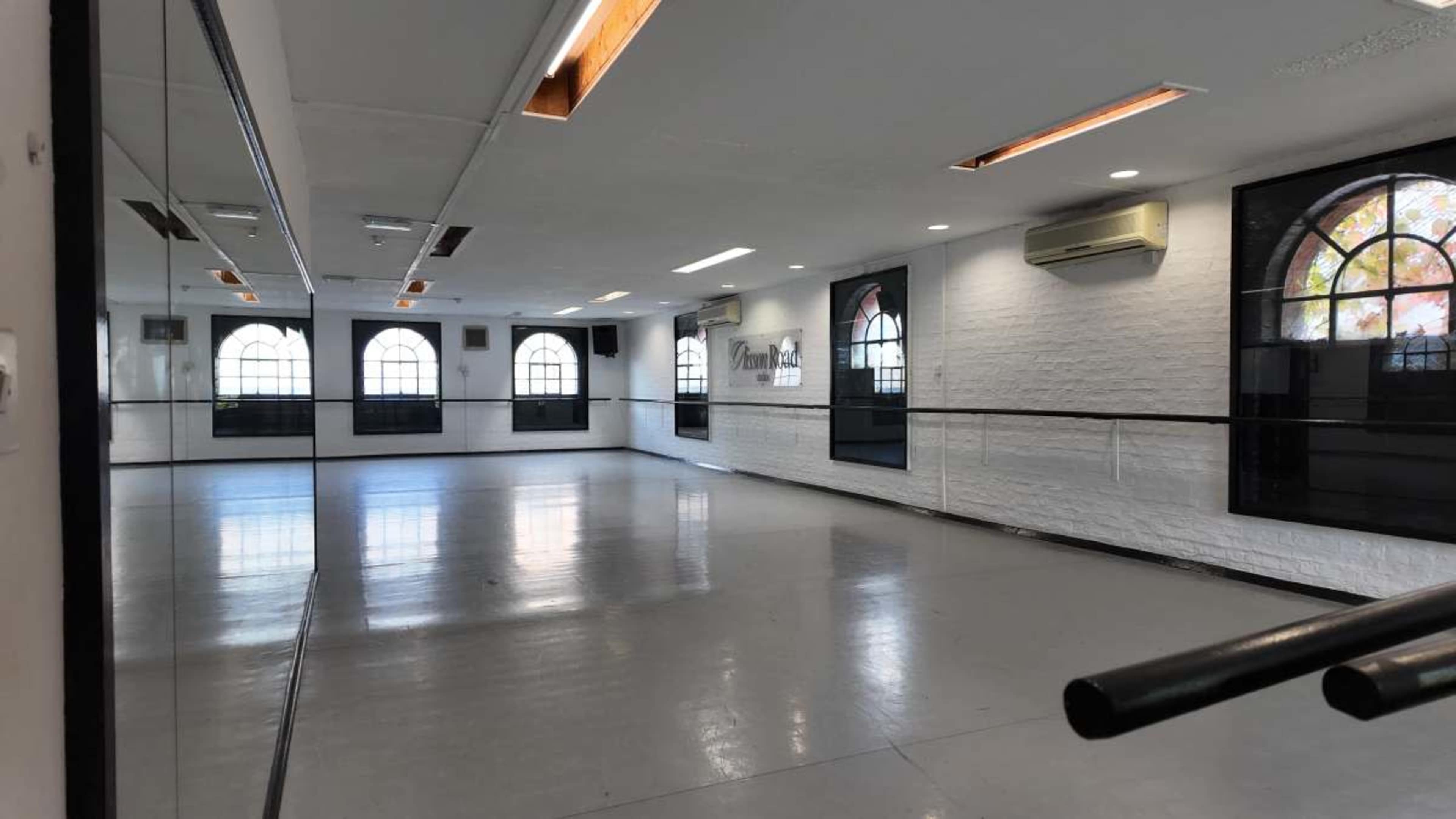 The image shows a spacious, empty dance studio with mirrors, wooden flooring, and large windows letting in natural light.