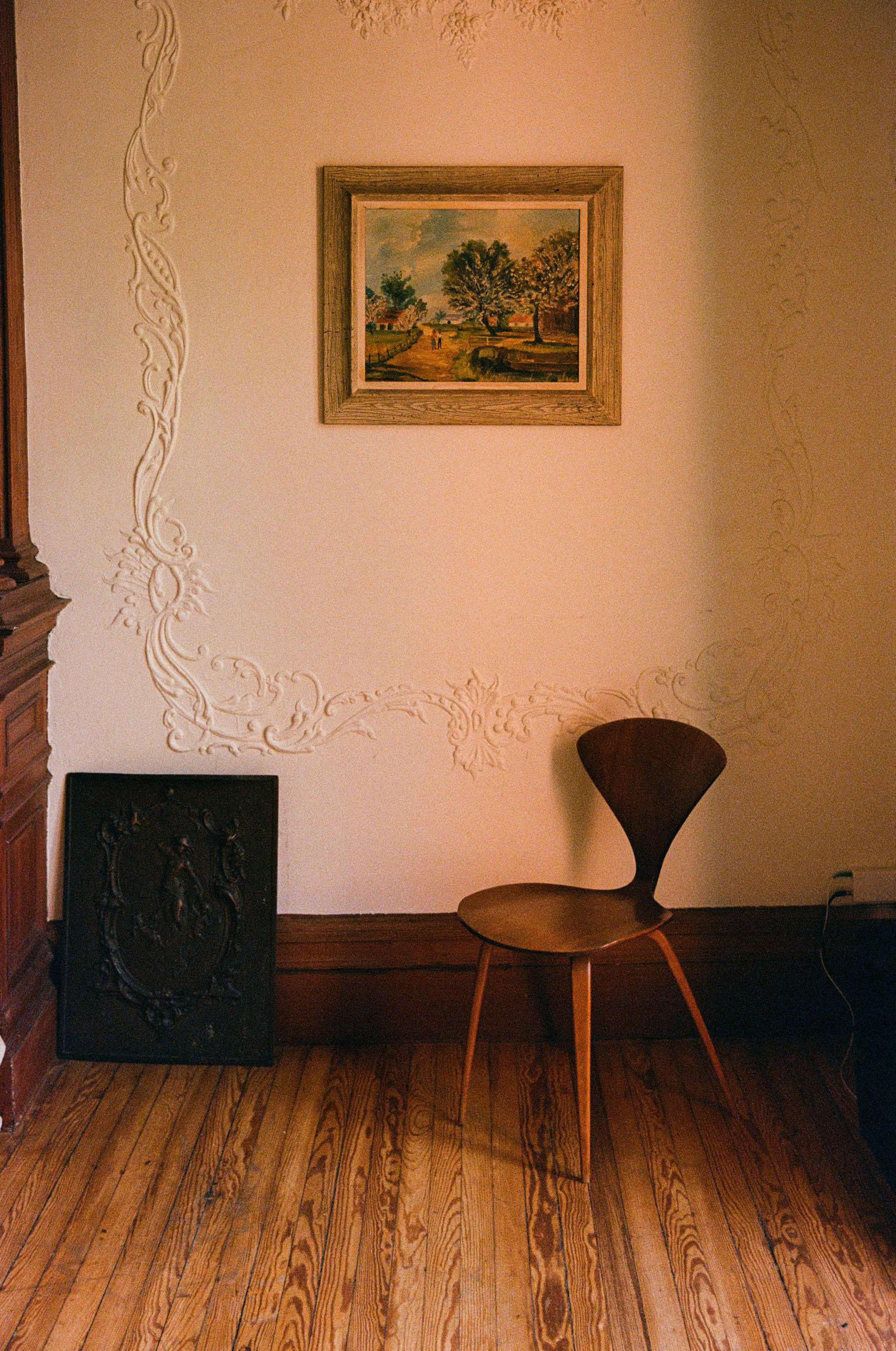 The image shows a minimalist corner of a room with a wooden chair positioned near a wall adorned with a painting and decorative molding.