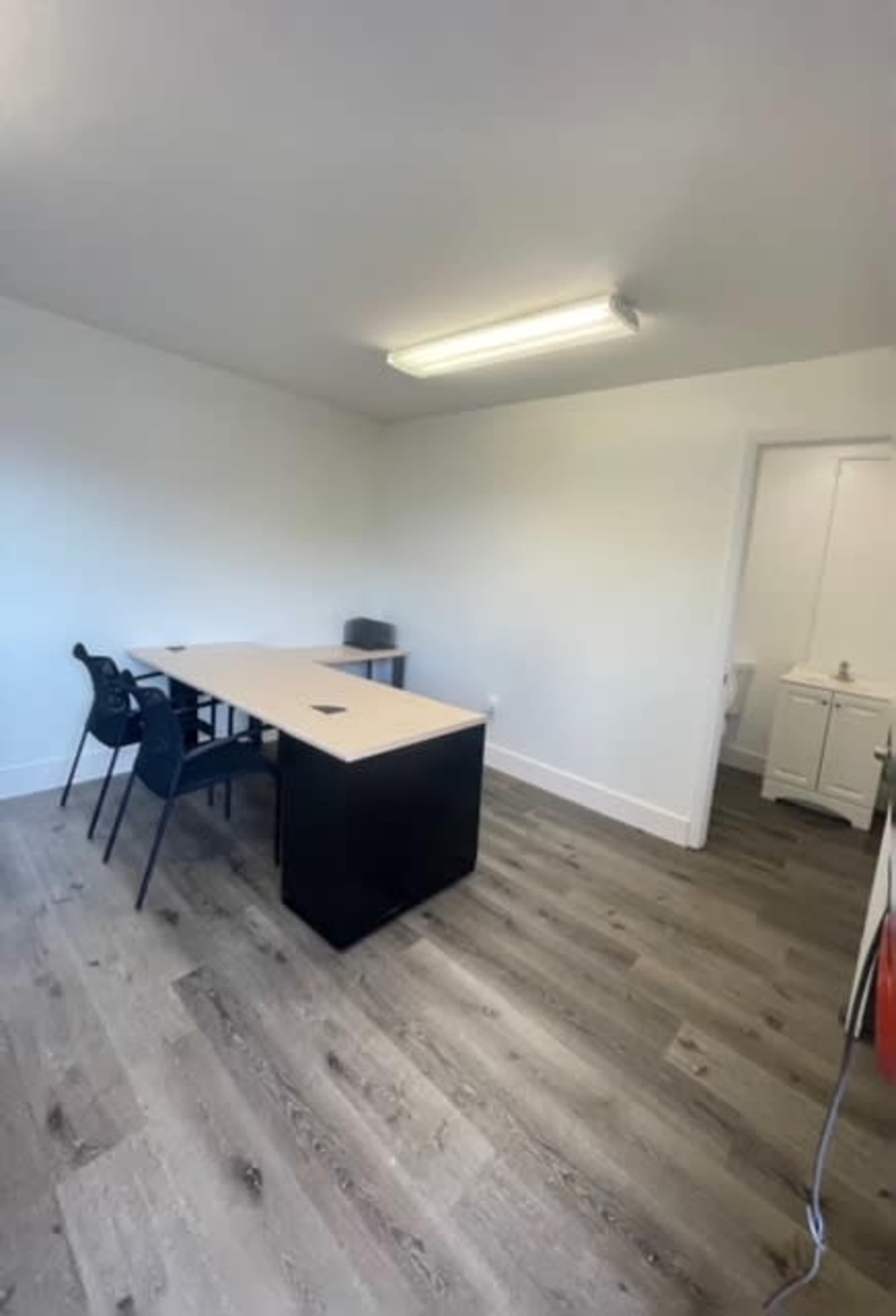 A sparsely furnished office features a long desk with two chairs and a door leading to a separate area.