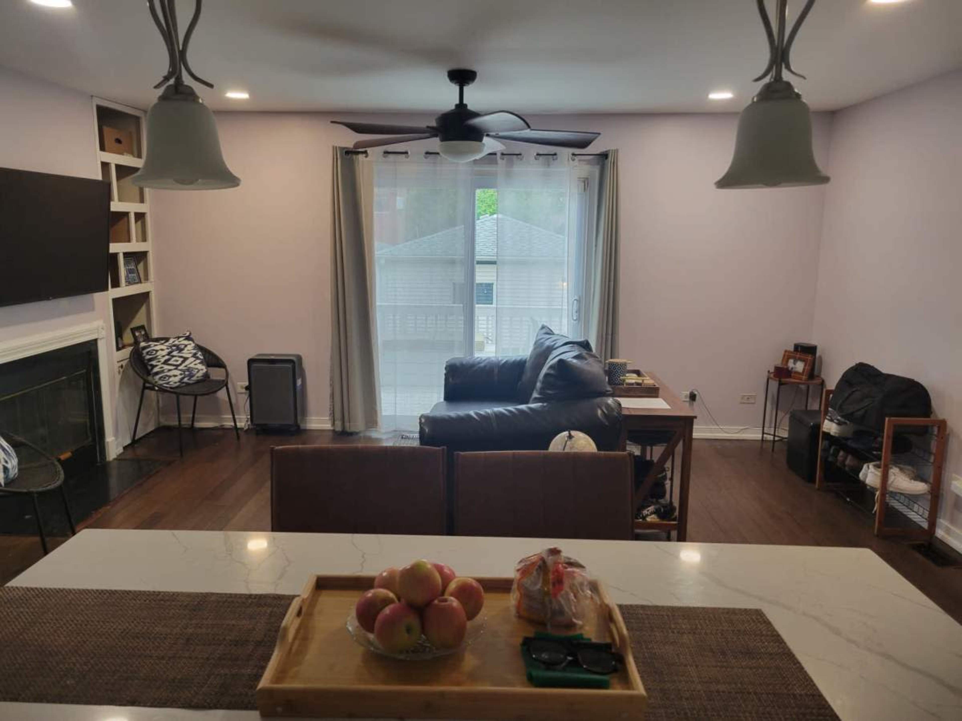 The image shows a living room and kitchen area with a leather couch, a coffee table, and large windows letting in natural light.