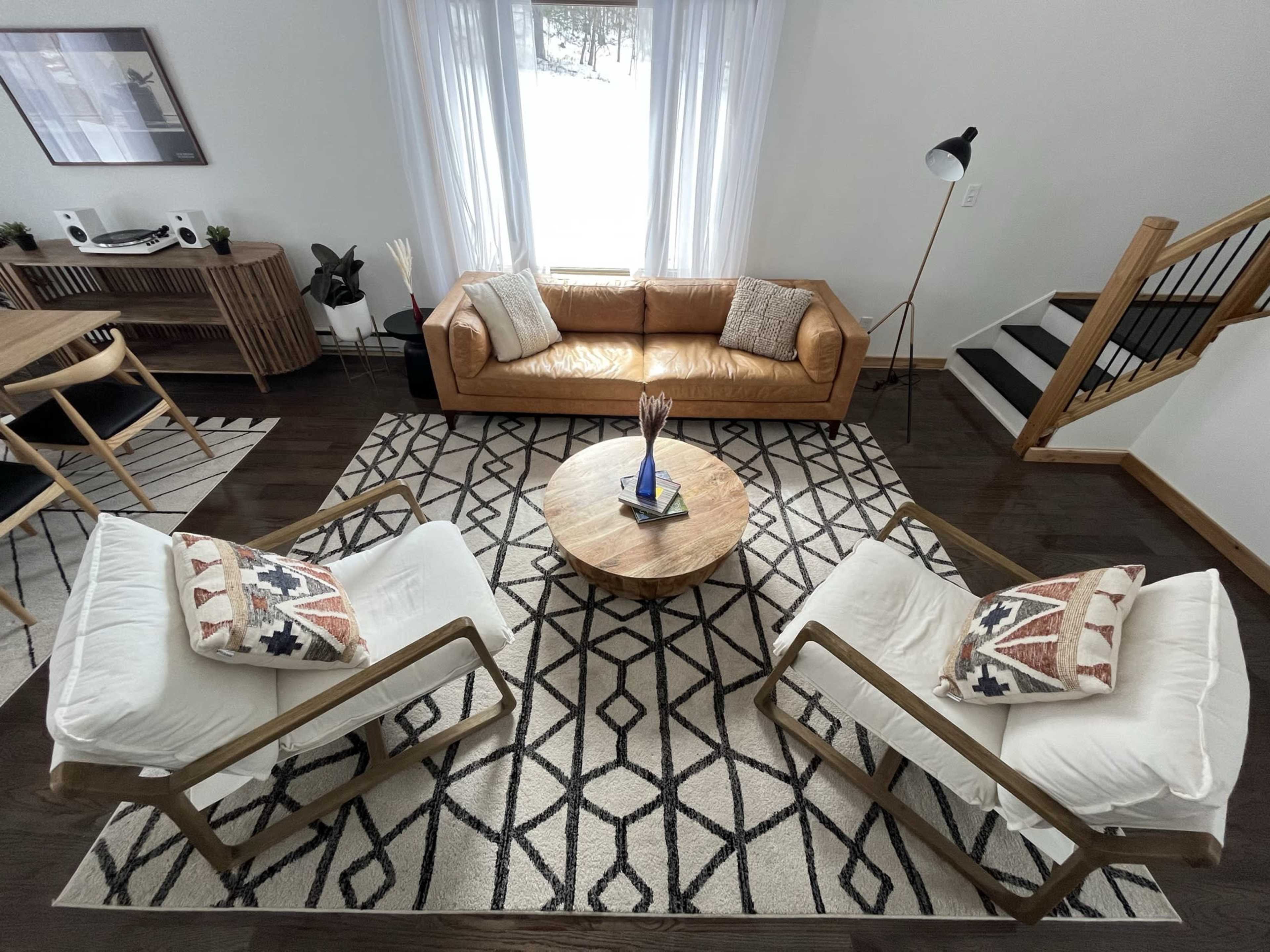 A modern living room with a leather sofa, two armchairs, a wooden coffee table, and a patterned area rug, illuminated by natural light from a large window.