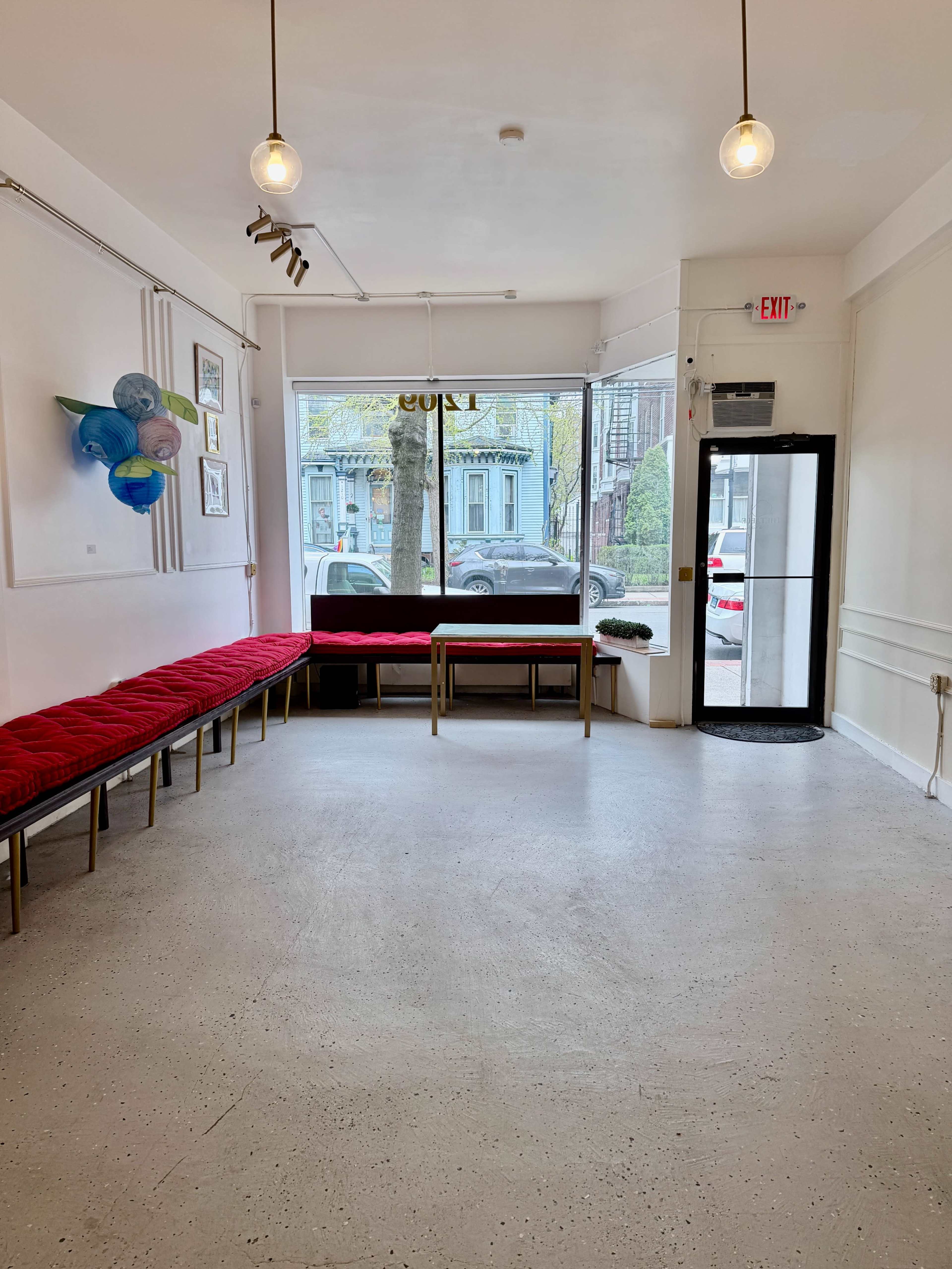 The image shows a bright, empty room with a red cushioned bench along one wall, a table in the center, and large windows facing the street.