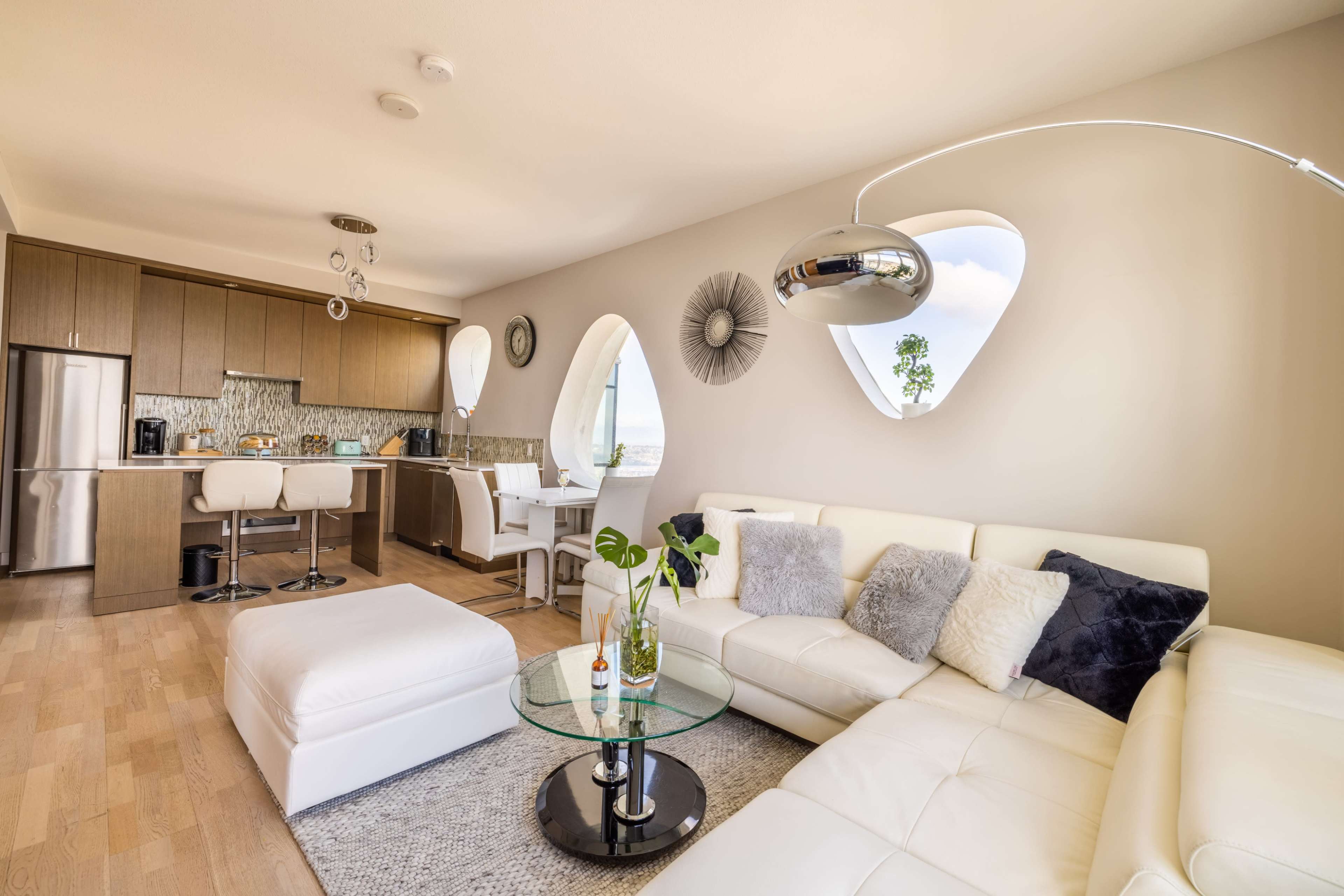 A modern living space with a white leather sofa, round glass coffee table, and a kitchen area with wooden cabinets and unique arched windows.