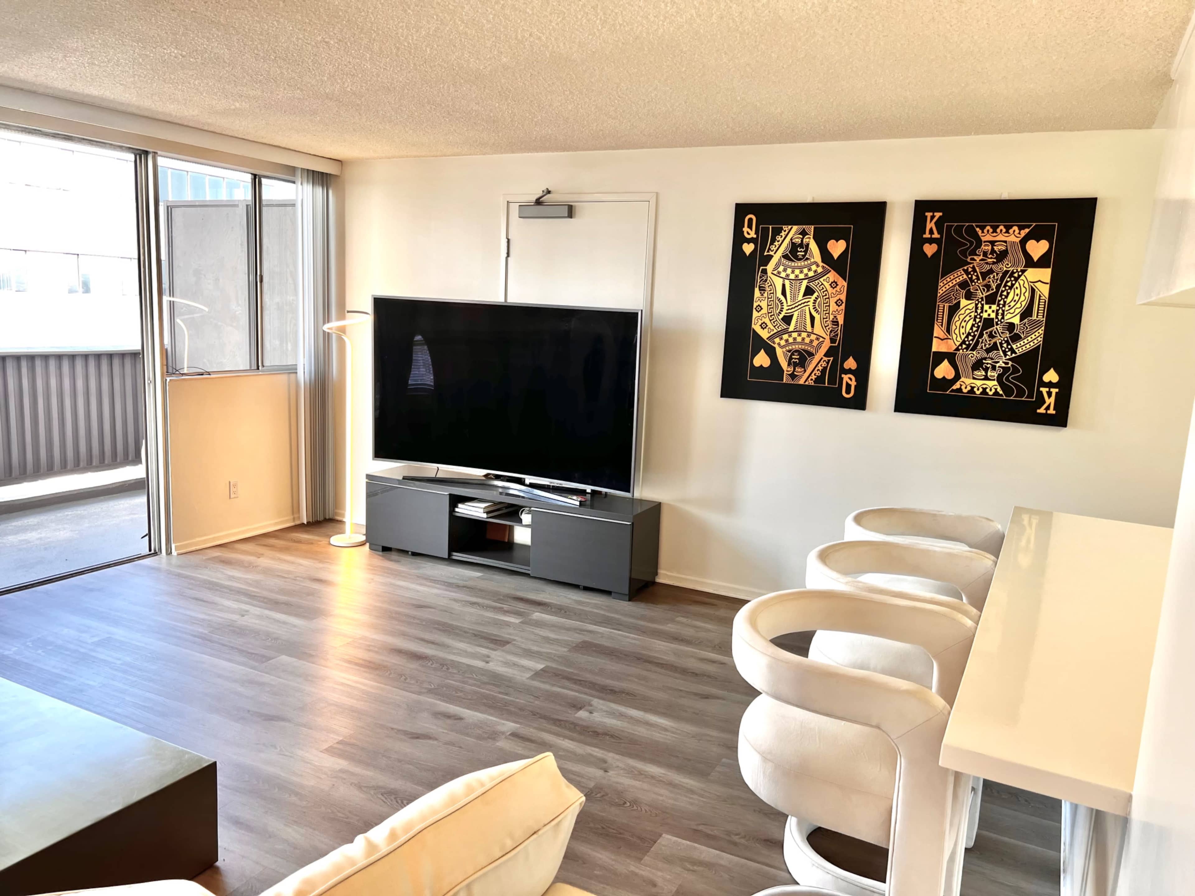 A well-lit living area features a large TV on a low cabinet, two framed playing card art pieces on the wall, and modern white chairs around a bar-style table.
