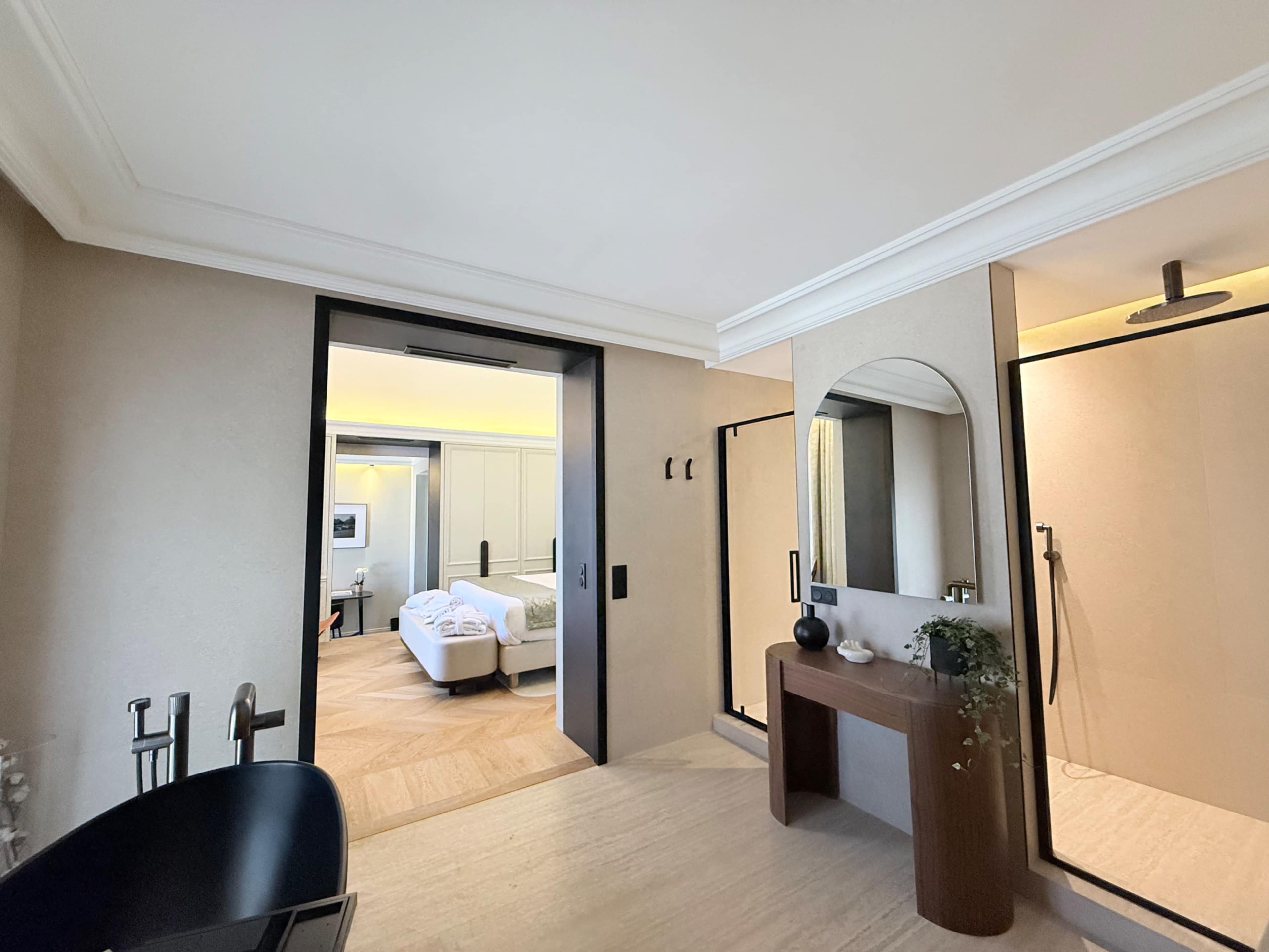 The image shows a modern bathroom with a sleek design, featuring a freestanding black bathtub, a stylish vanity with a round mirror, and an open doorway leading to a well-lit bedroom.
