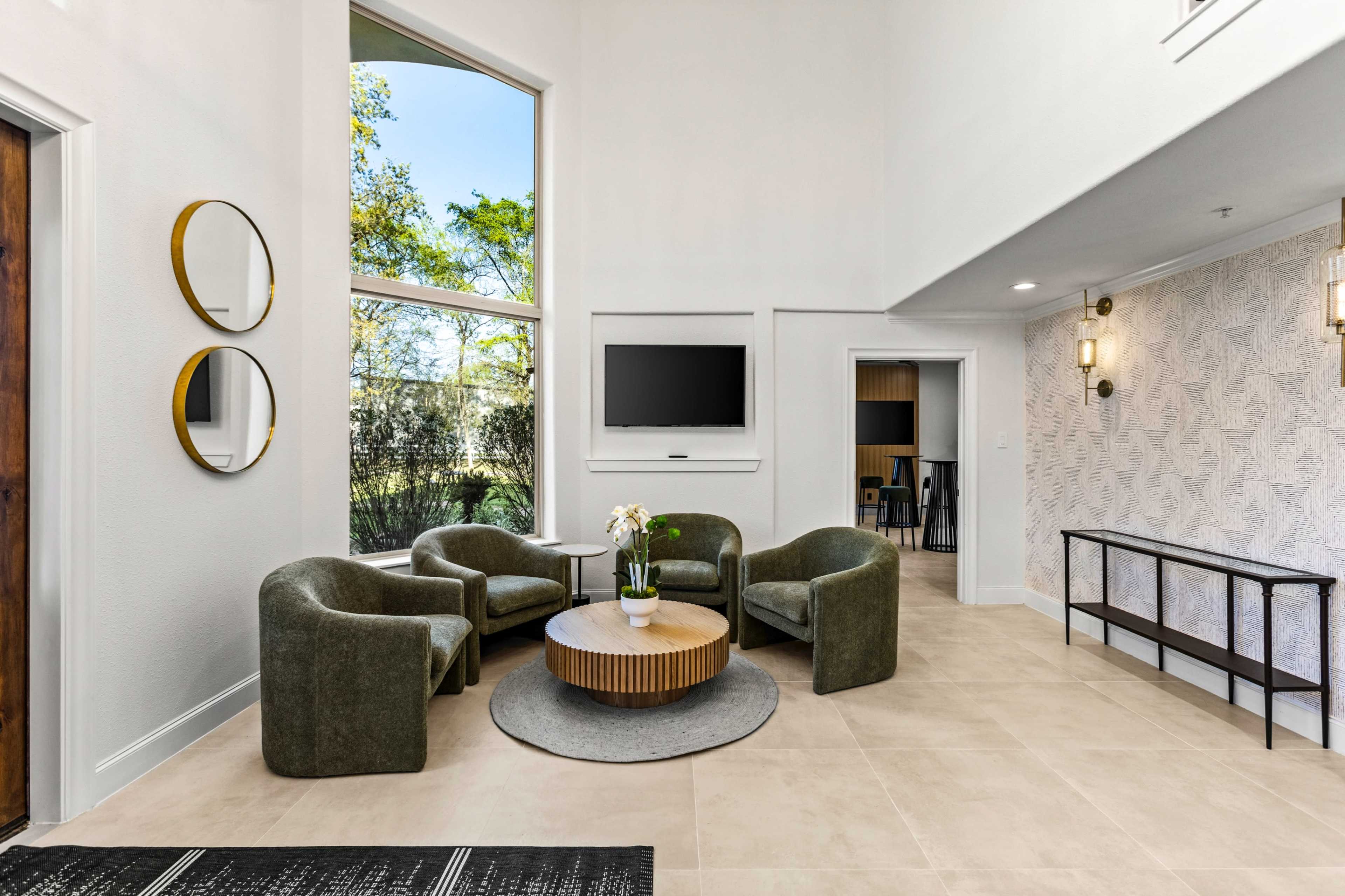 The image shows a modern living area with four upholstered chairs arranged around a circular table, a television mounted on the wall, and large windows allowing natural light to enter.
