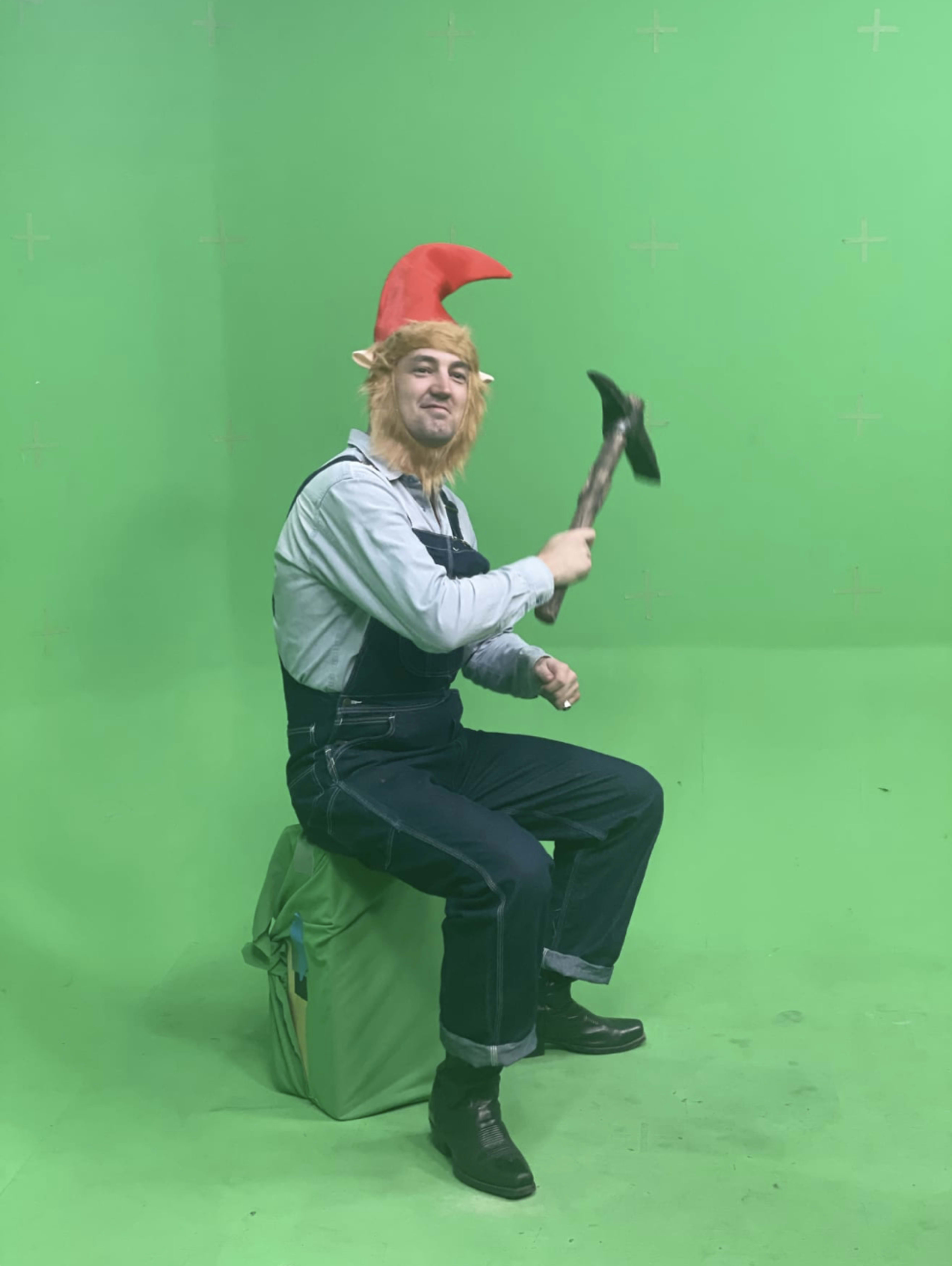 A person wearing a gnome costume, complete with a pointed hat and holding a hammer, sits on a green box in front of a green screen.