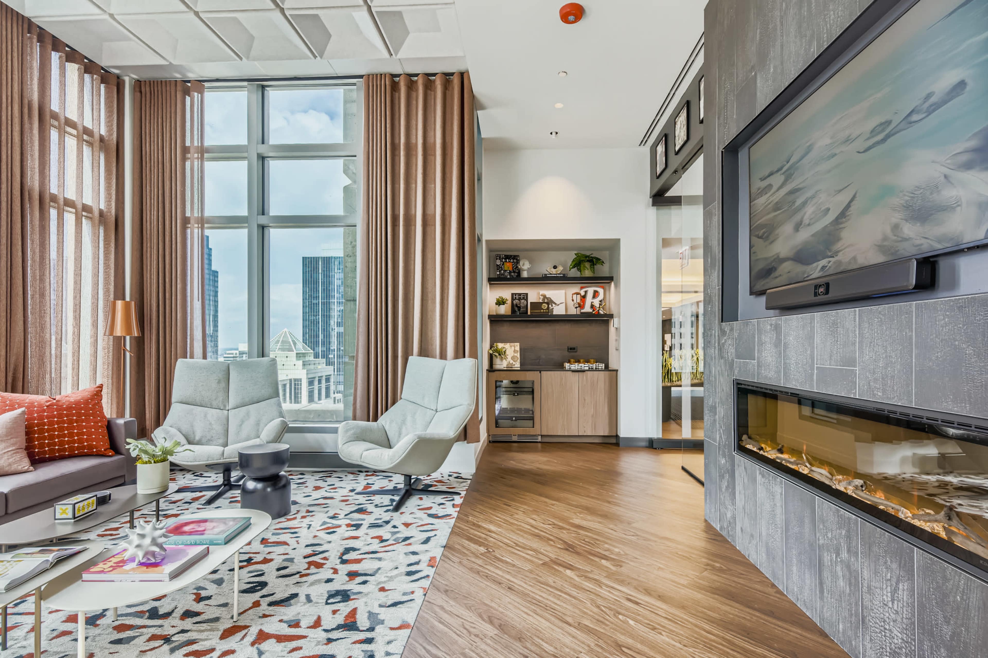 The image shows a modern living room with large windows, two light-colored armchairs, a sofa, and a decorative area rug, accompanied by a flat-screen TV mounted on a textured wall.