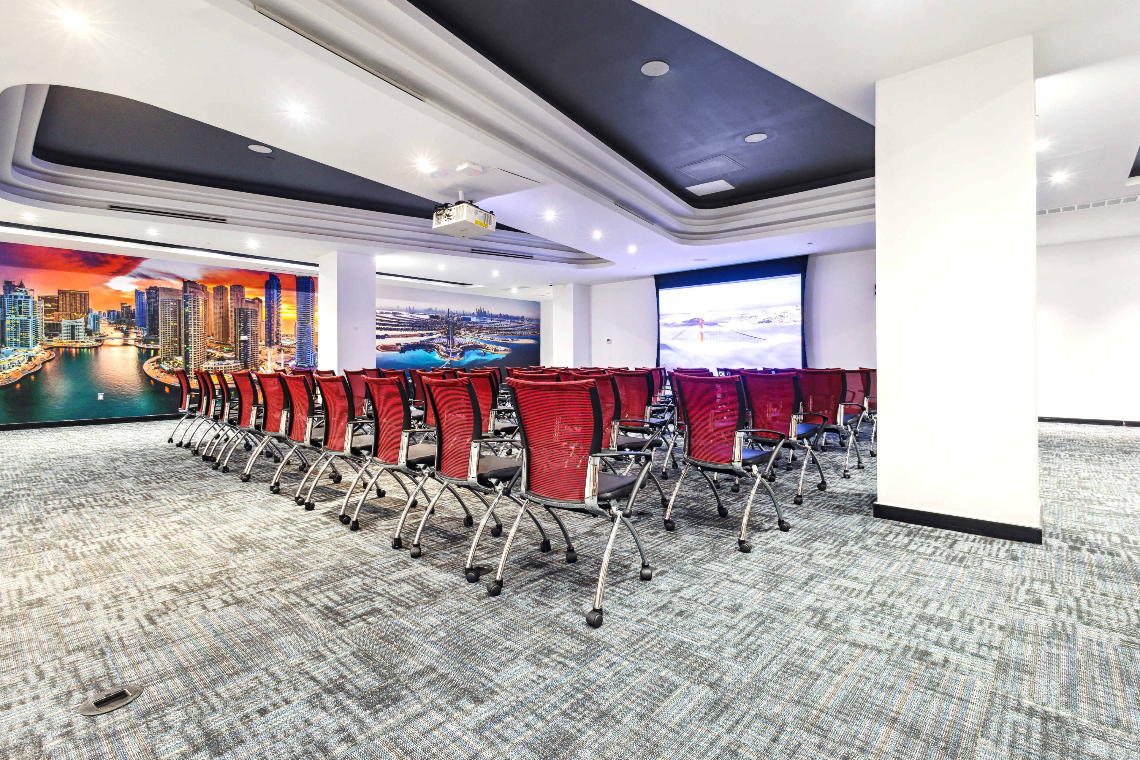 The image shows a conference room arranged with rows of red chairs facing a large projection screen, with a cityscape mural on the side wall.