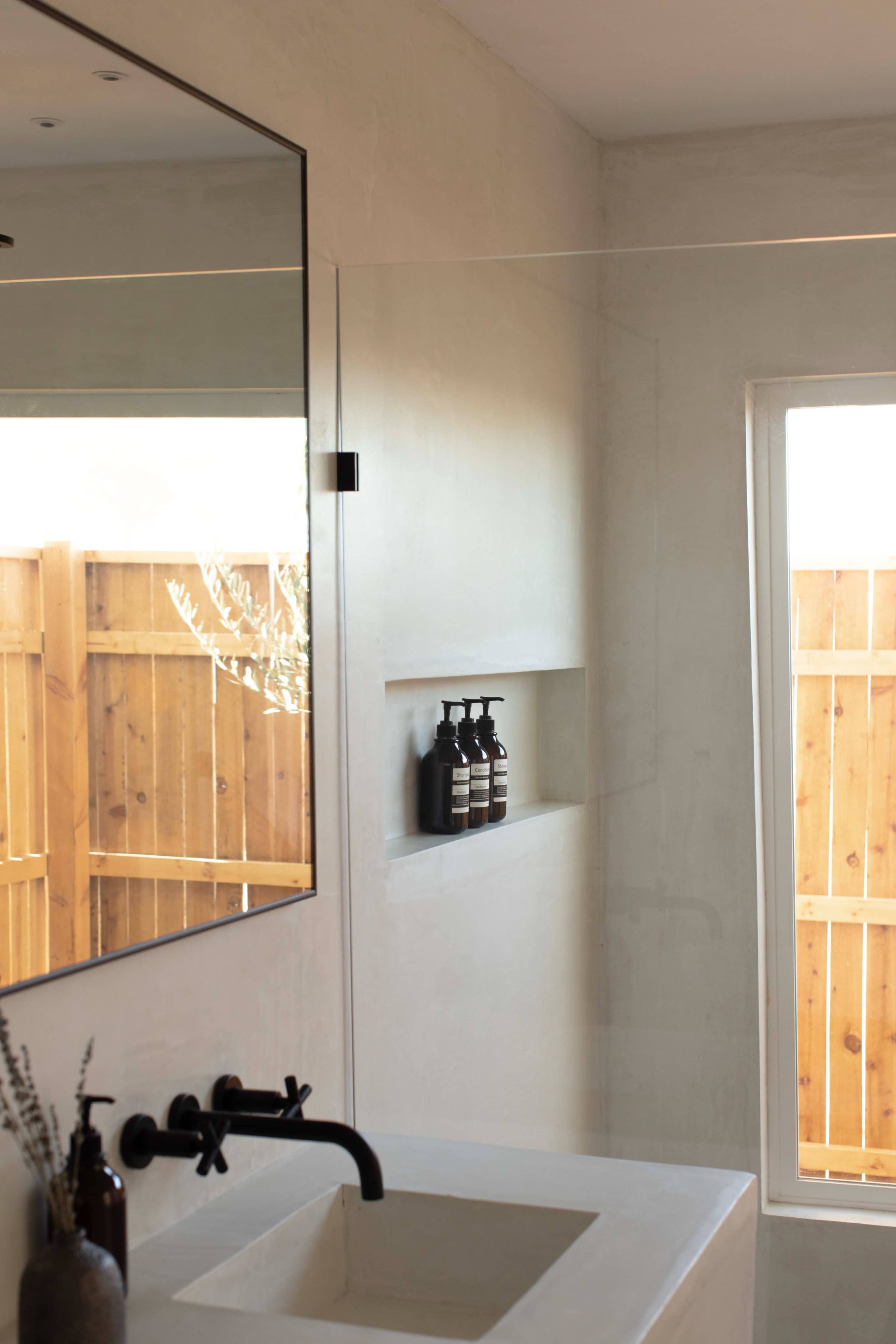 A modern bathroom features a large mirror, a minimalist sink, and a niche displaying dark bottles, with a wooden fence visible through the window.