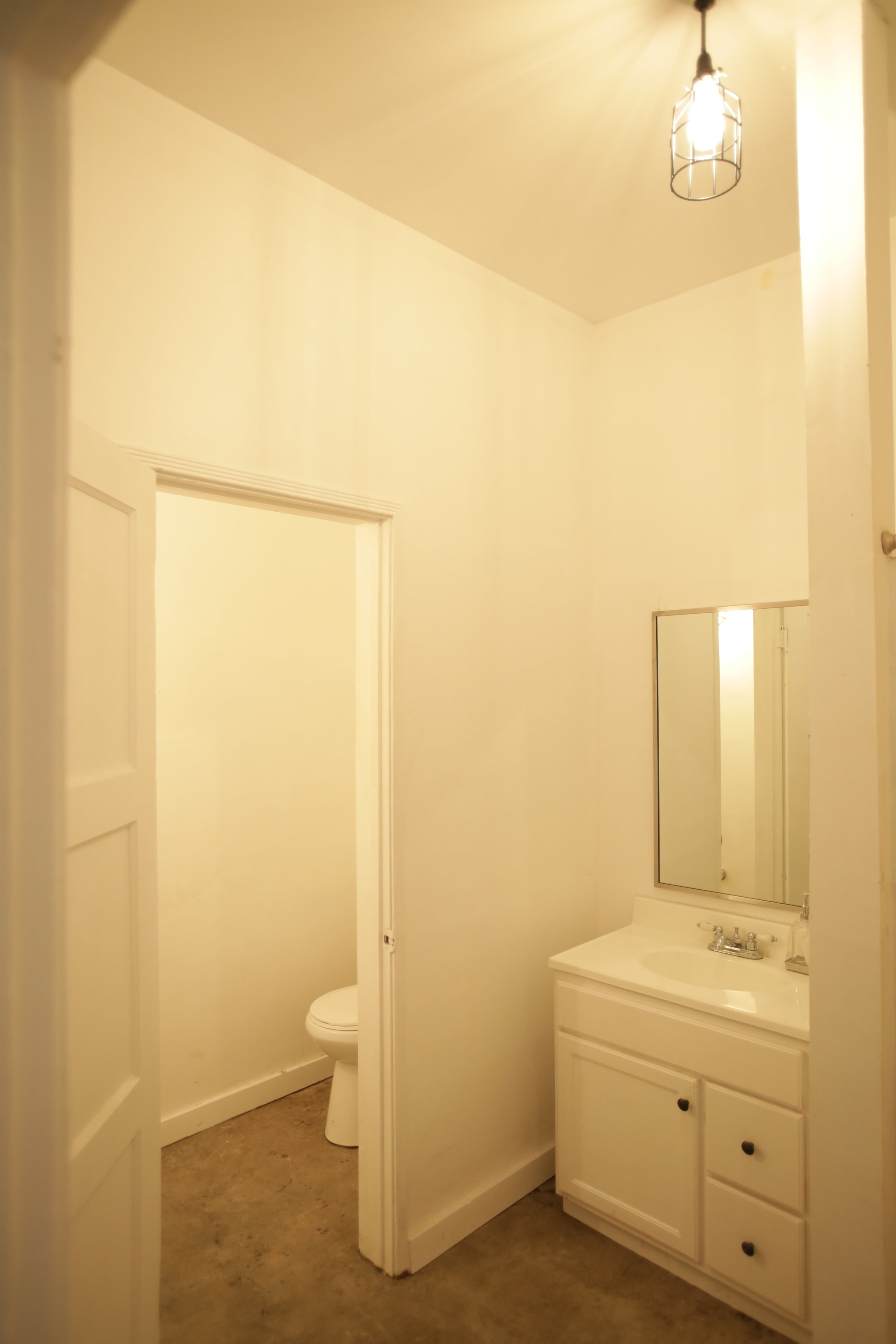 A small bathroom features a sink with a mirror, a door leading to a toilet, and a light fixture hanging from the ceiling.