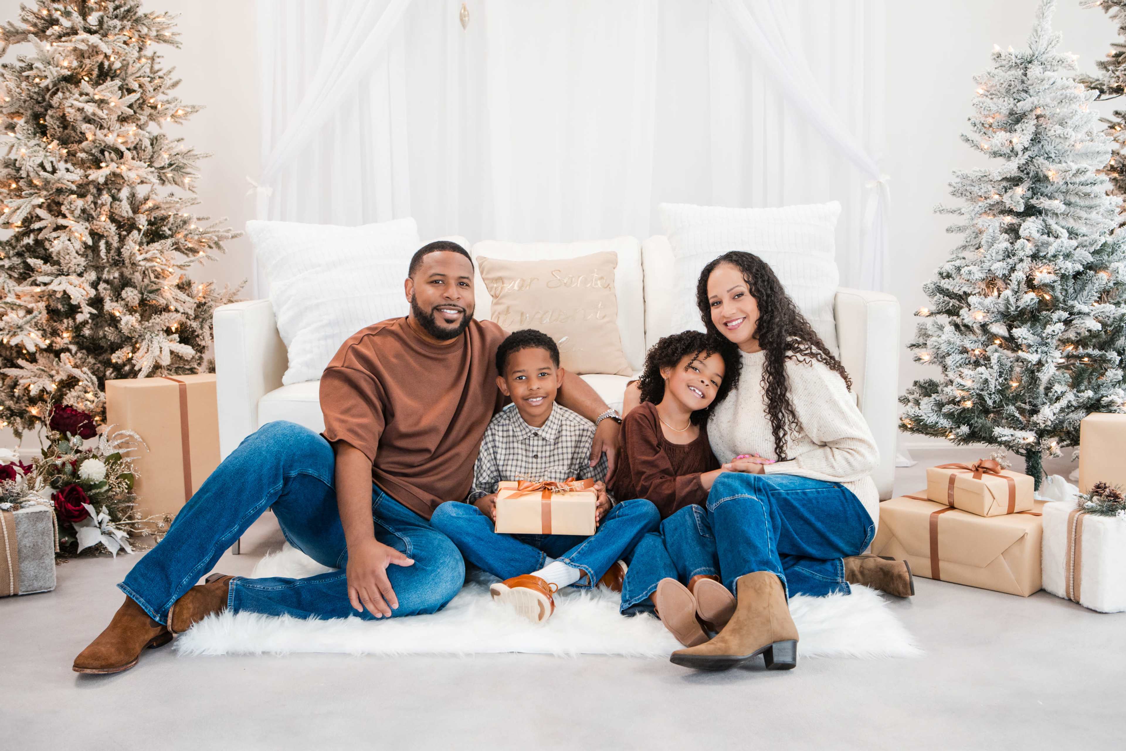 A family of four sits together on a white rug in a festive living room decorated with Christmas trees and wrapped gifts.