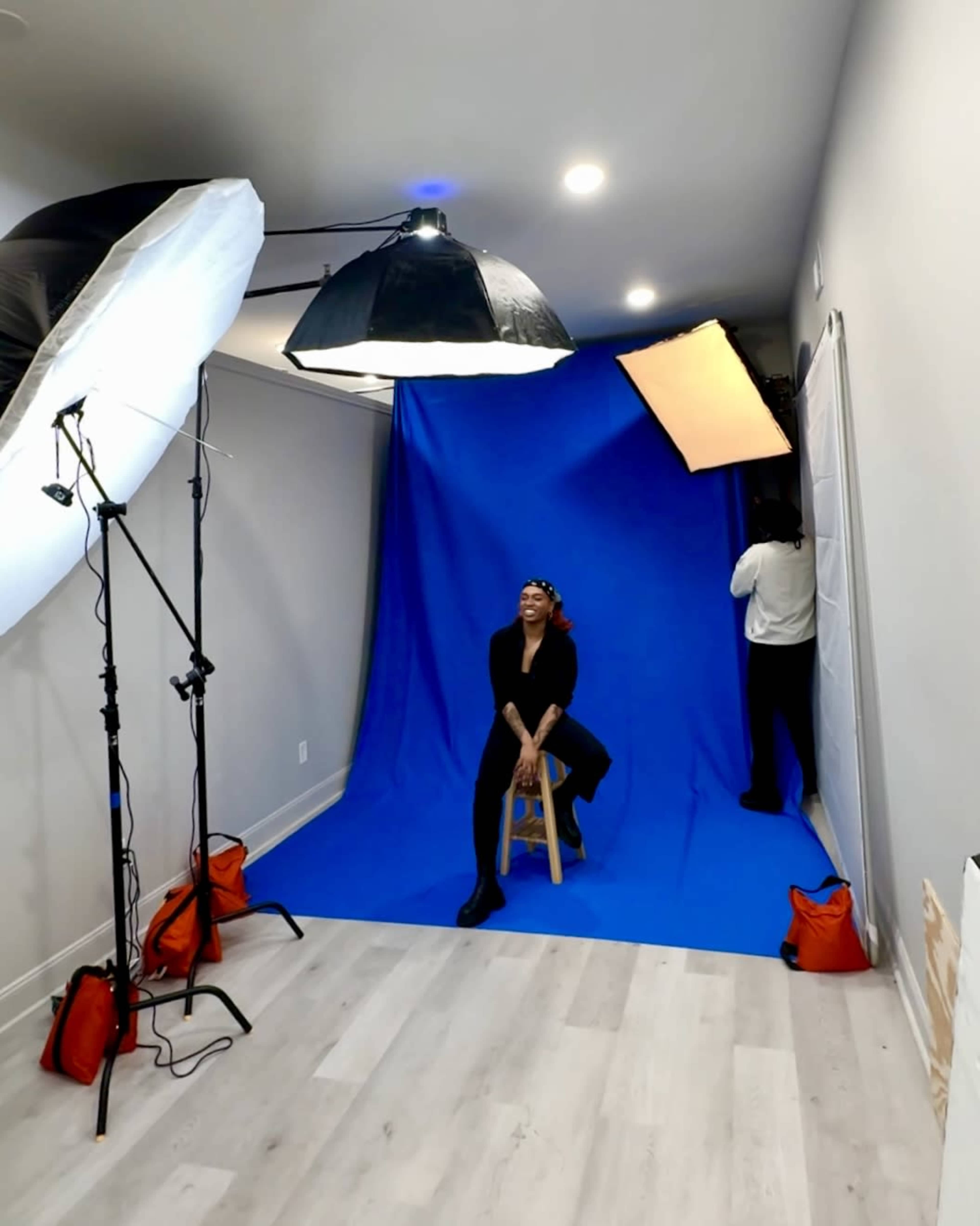 A woman sits on a stool in front of a bright blue backdrop, surrounded by studio lighting equipment.