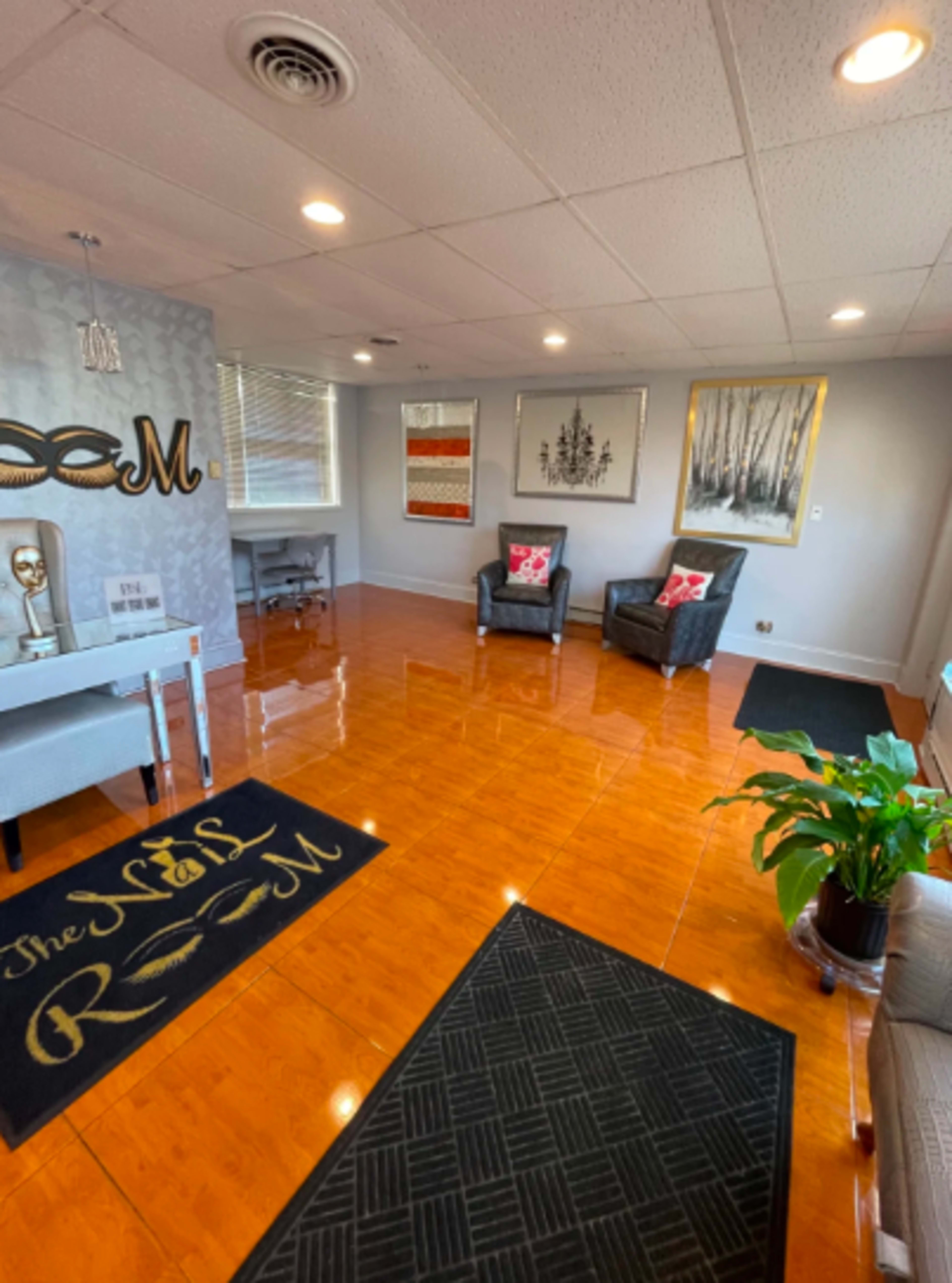 The image shows a well-lit nail salon waiting area with a polished wooden floor, two chairs with decorative pillows, and artwork on the walls.