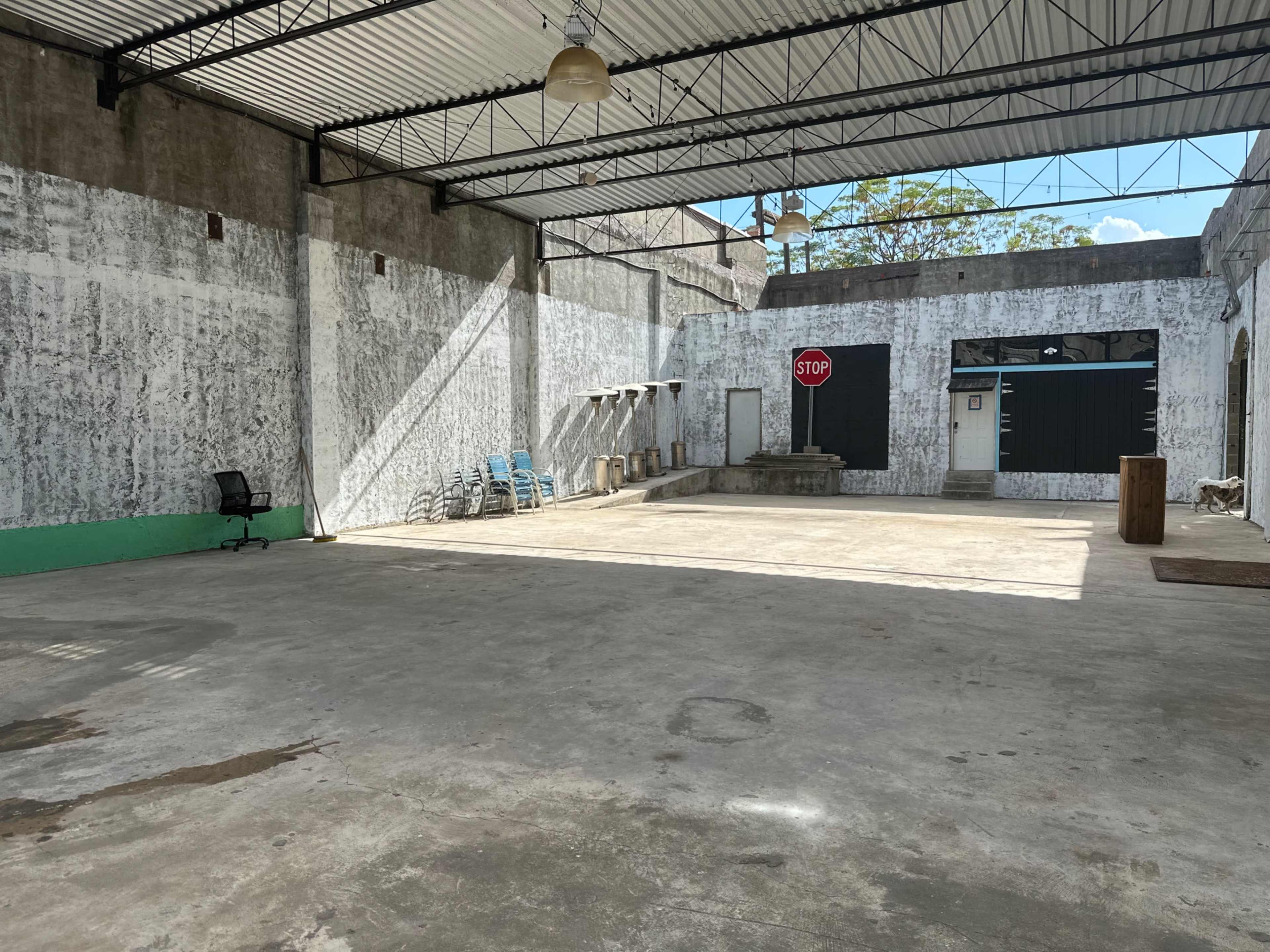 The image shows a large, empty industrial space with concrete floors, a few chairs stacked against the wall, and a stop sign in the background.