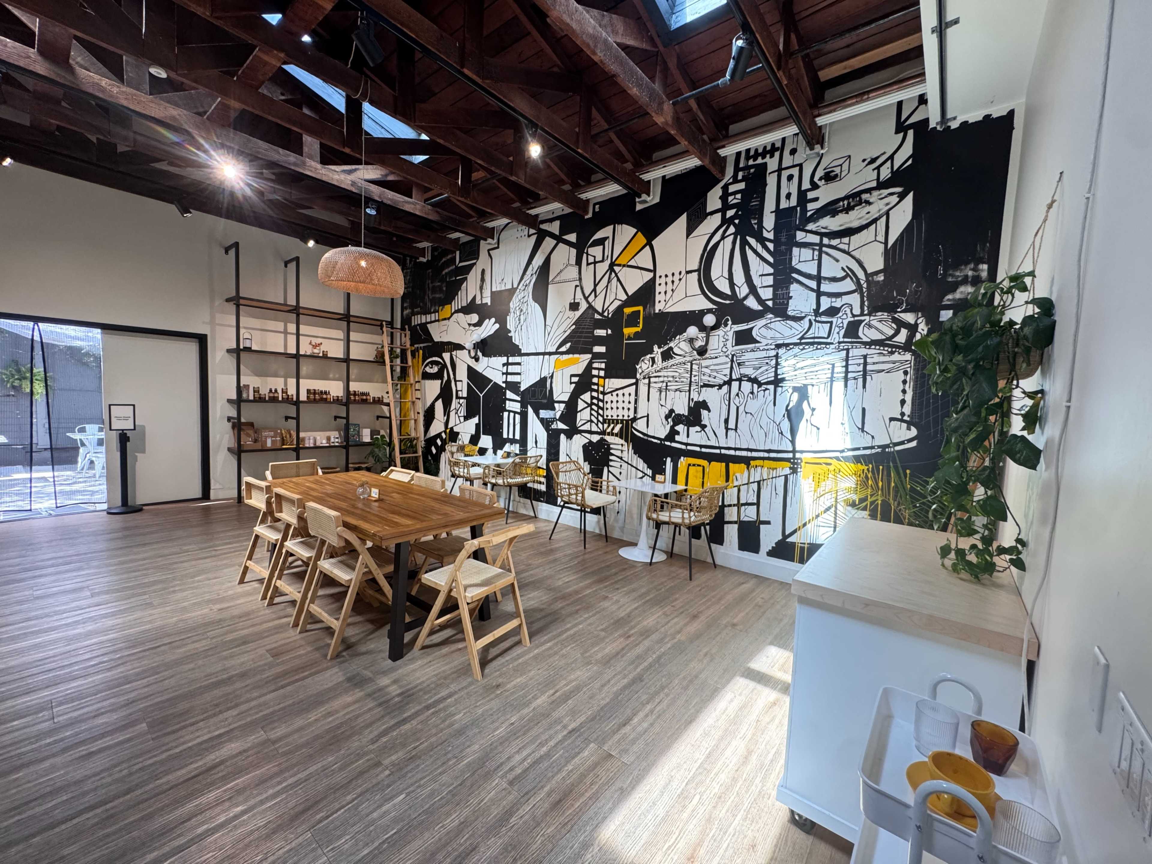 The image shows a spacious, modern dining area featuring a large wooden table and a vibrant mural on one wall, accompanied by a few plants and minimalist furniture.