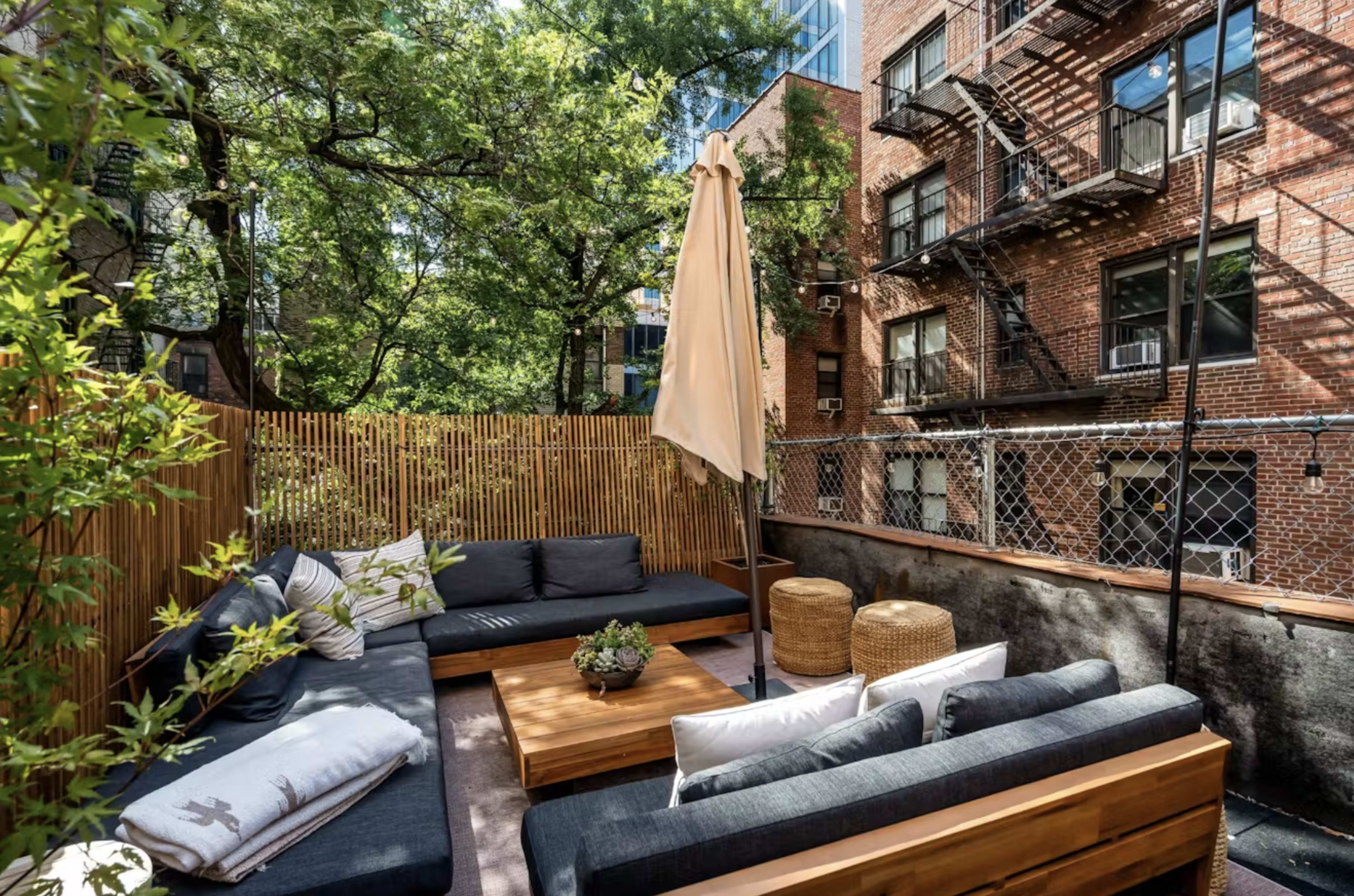 The image shows a small outdoor patio area furnished with a dark couch, wooden coffee table, and two straw stools, surrounded by greenery and brick buildings.