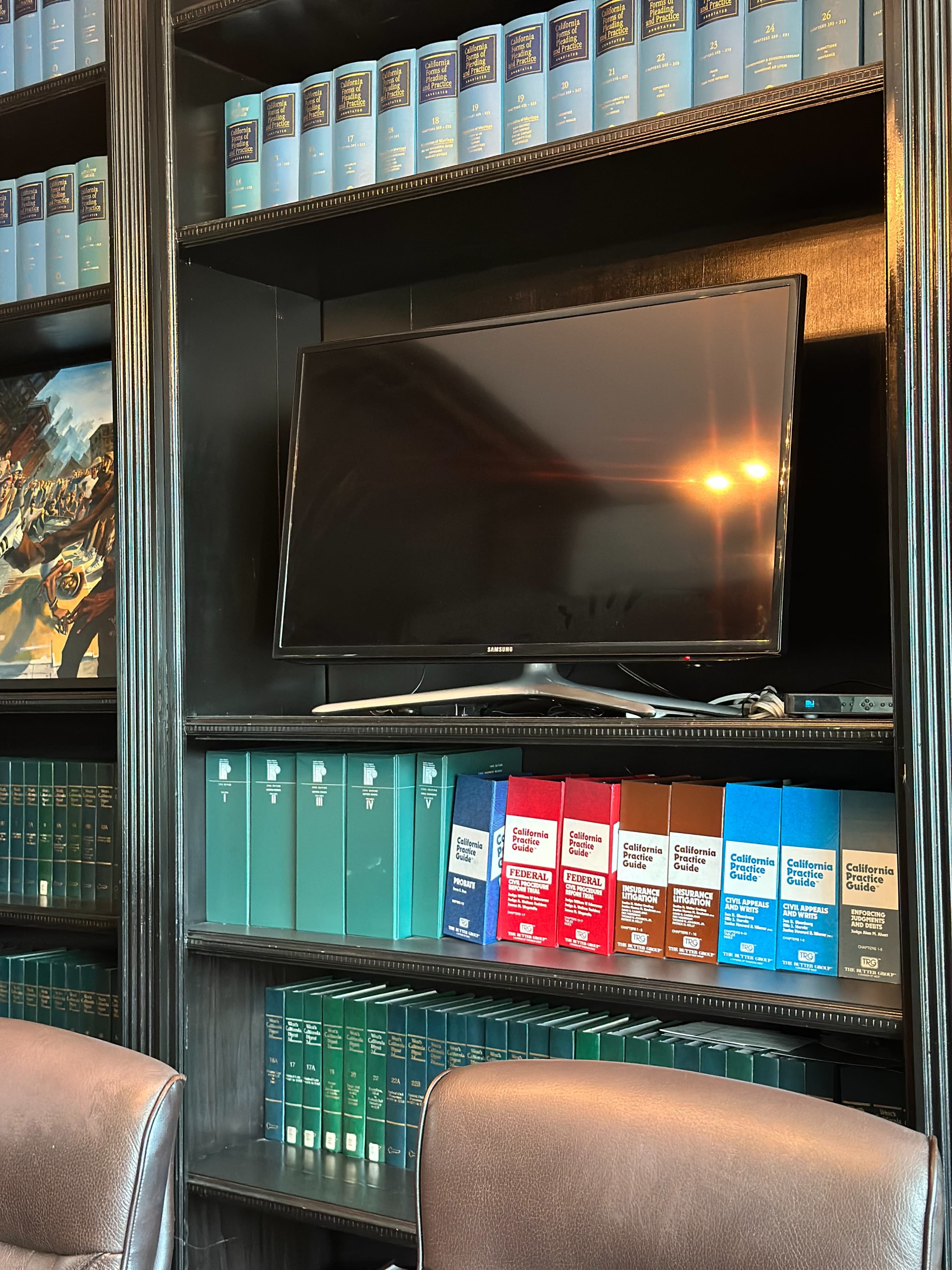 The image shows a bookshelf filled with various legal books and a television mounted above them in a room with brown leather chairs.