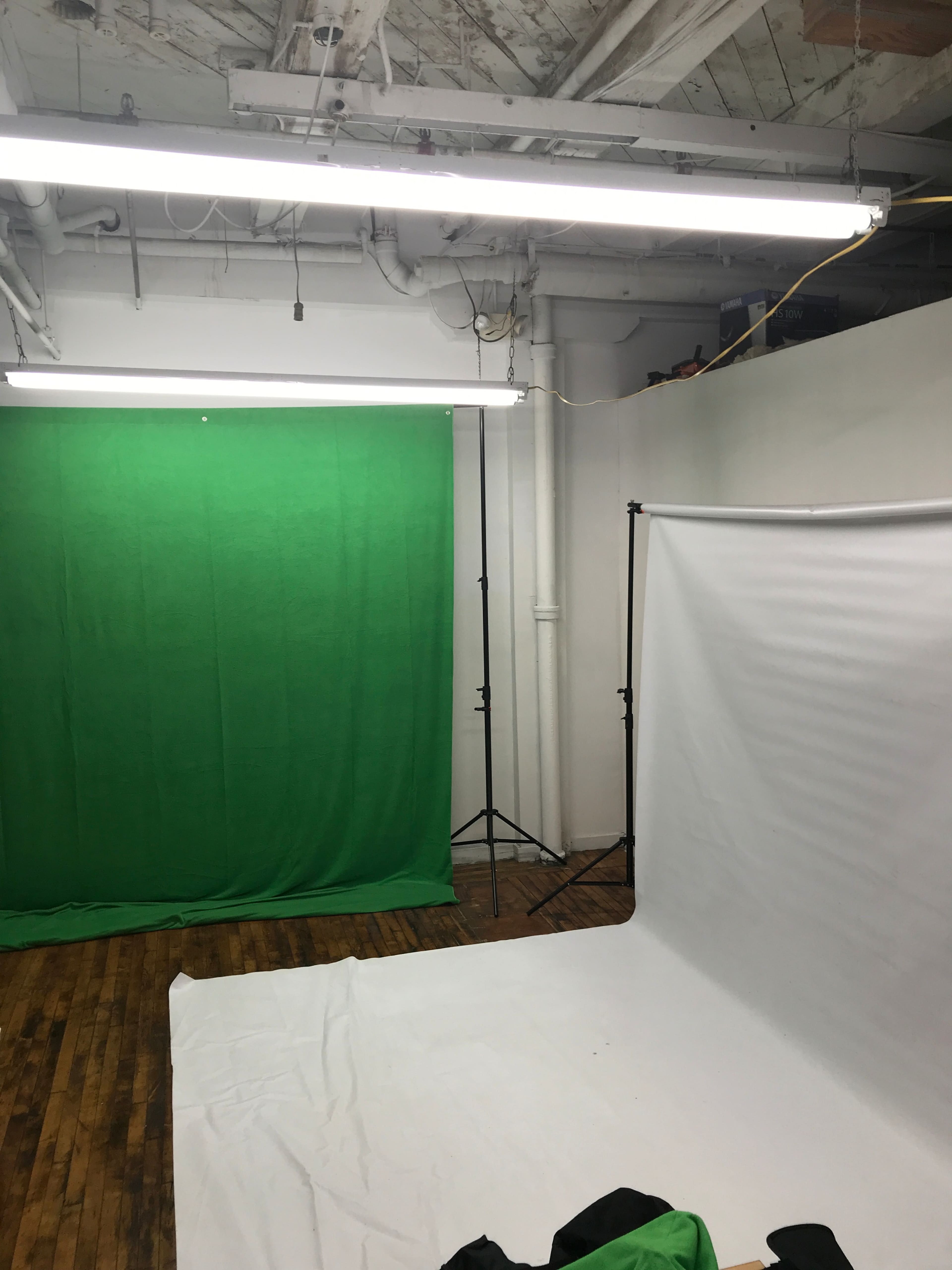 A photography studio features a green backdrop on one side and a white backdrop on the other, with a wooden floor and lighting fixtures above.
