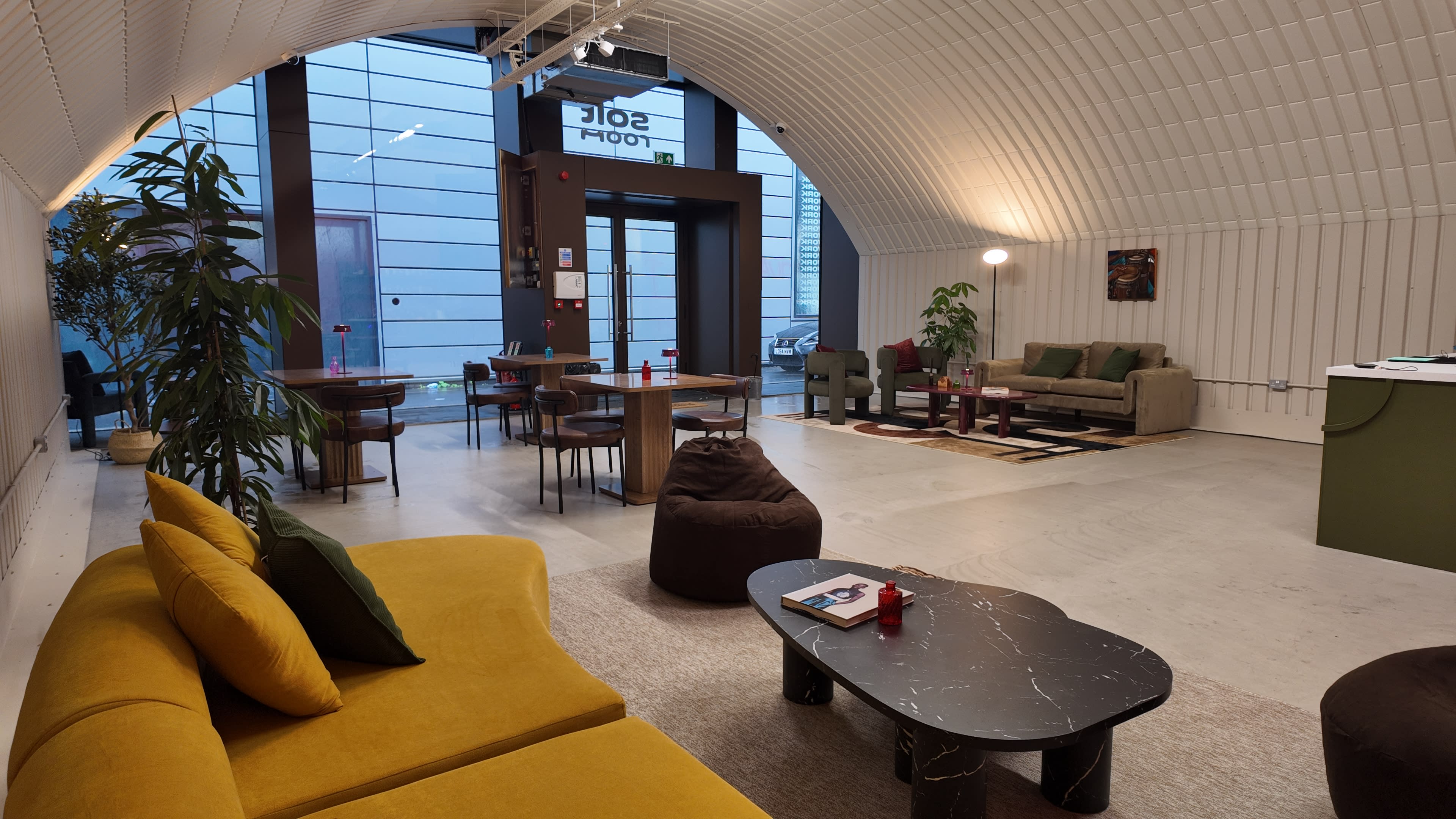 The image shows a modern, spacious lounge area with yellow couches, a black coffee table, and various seating arrangements, illuminated by natural light through large windows.