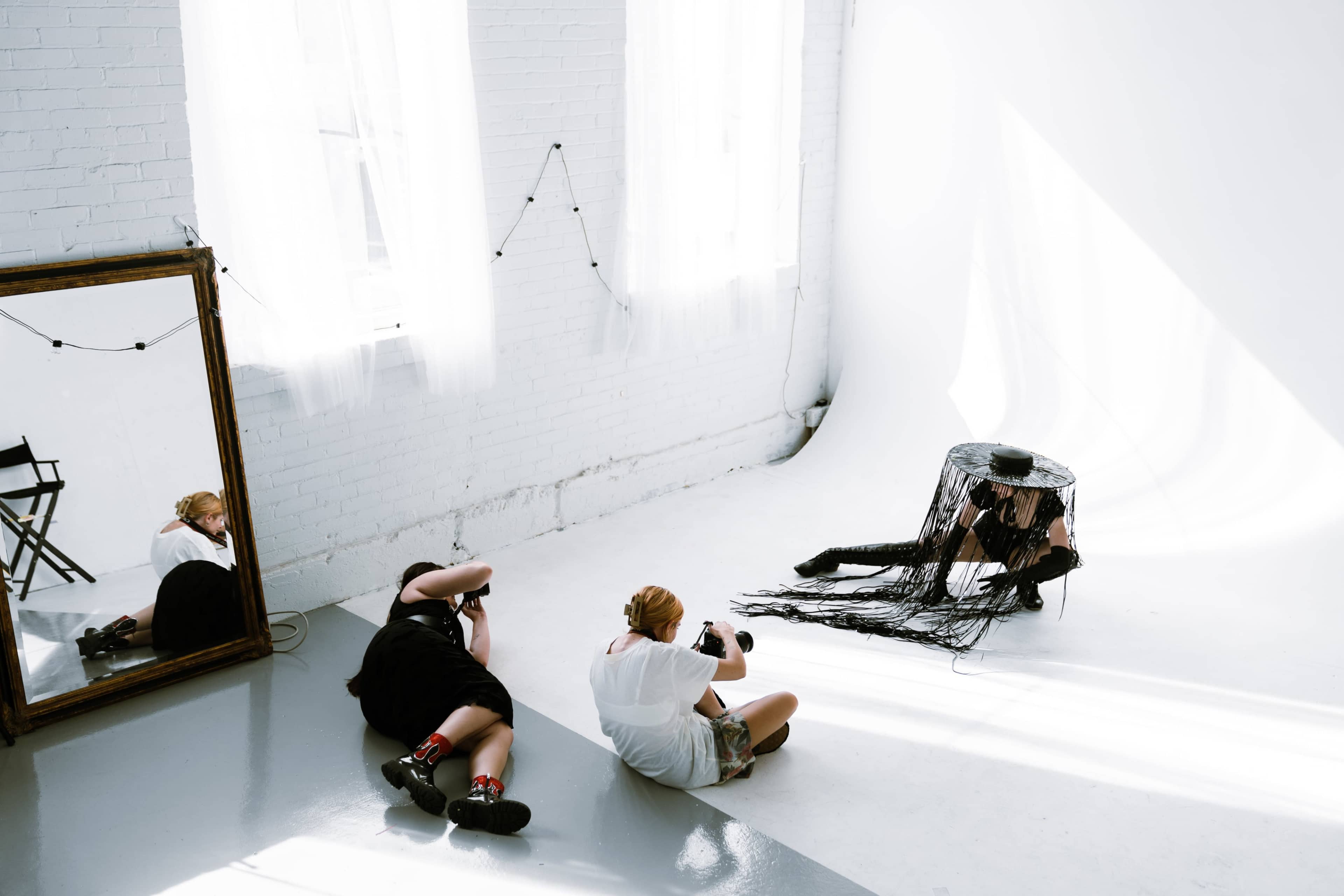 Two photographers capture images of a model posing under a fringed black hat in a bright, minimalist studio with a large mirror.