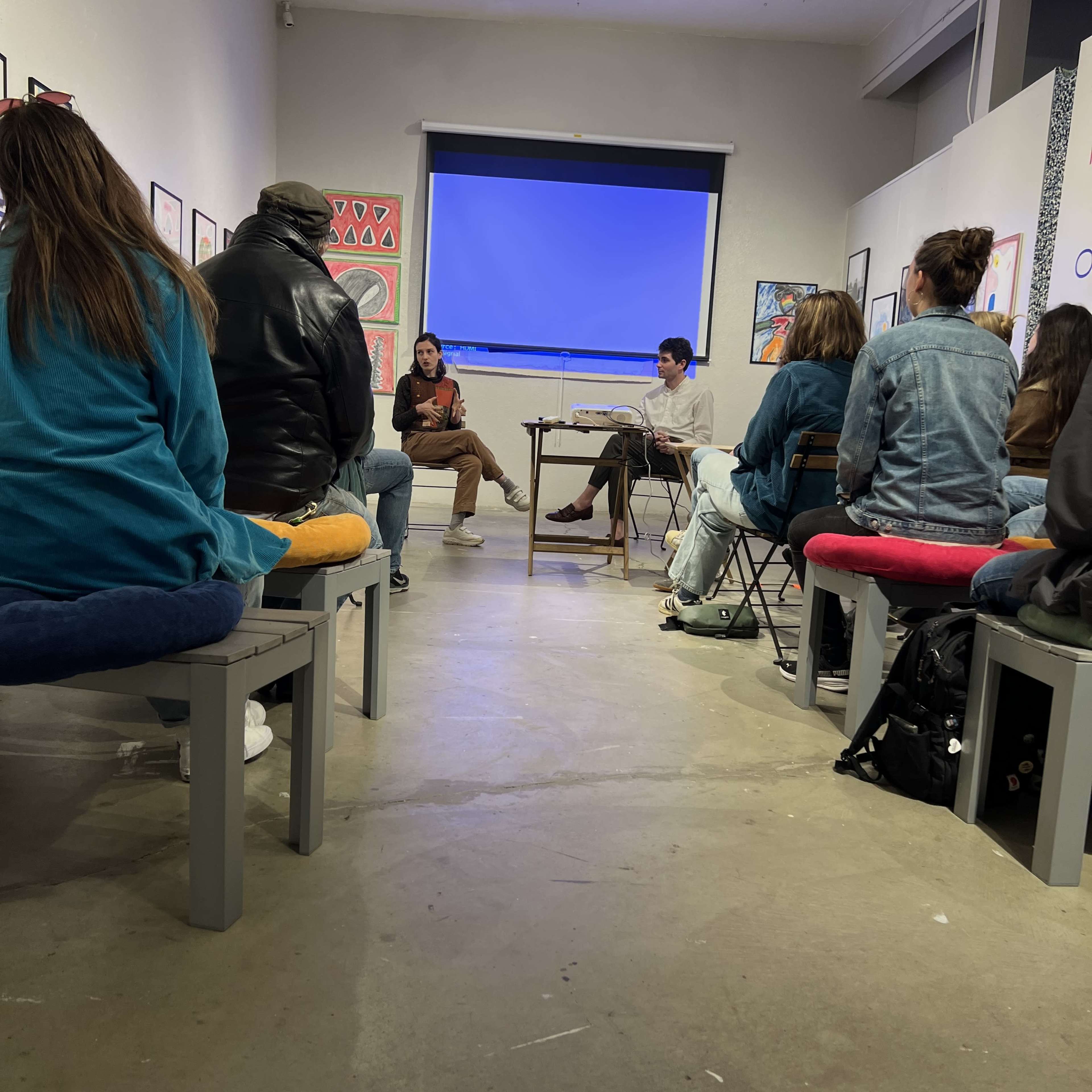 A group of people is seated on benches in a gallery, attentively listening to two individuals presenting at the front of the room.