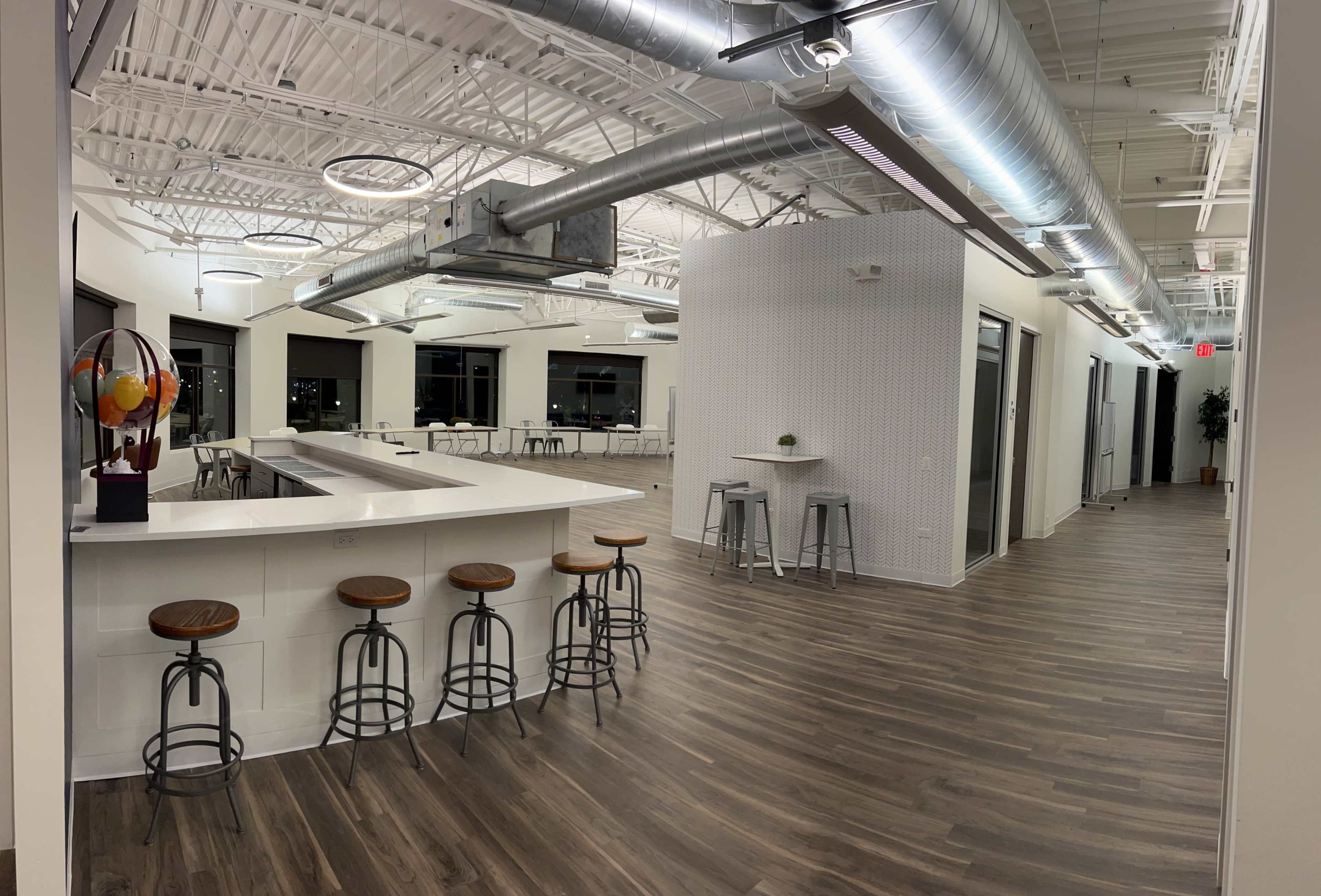 A spacious, modern interior with a kitchen area featuring a bar and stools, alongside an open seating space with tables in a well-lit environment.