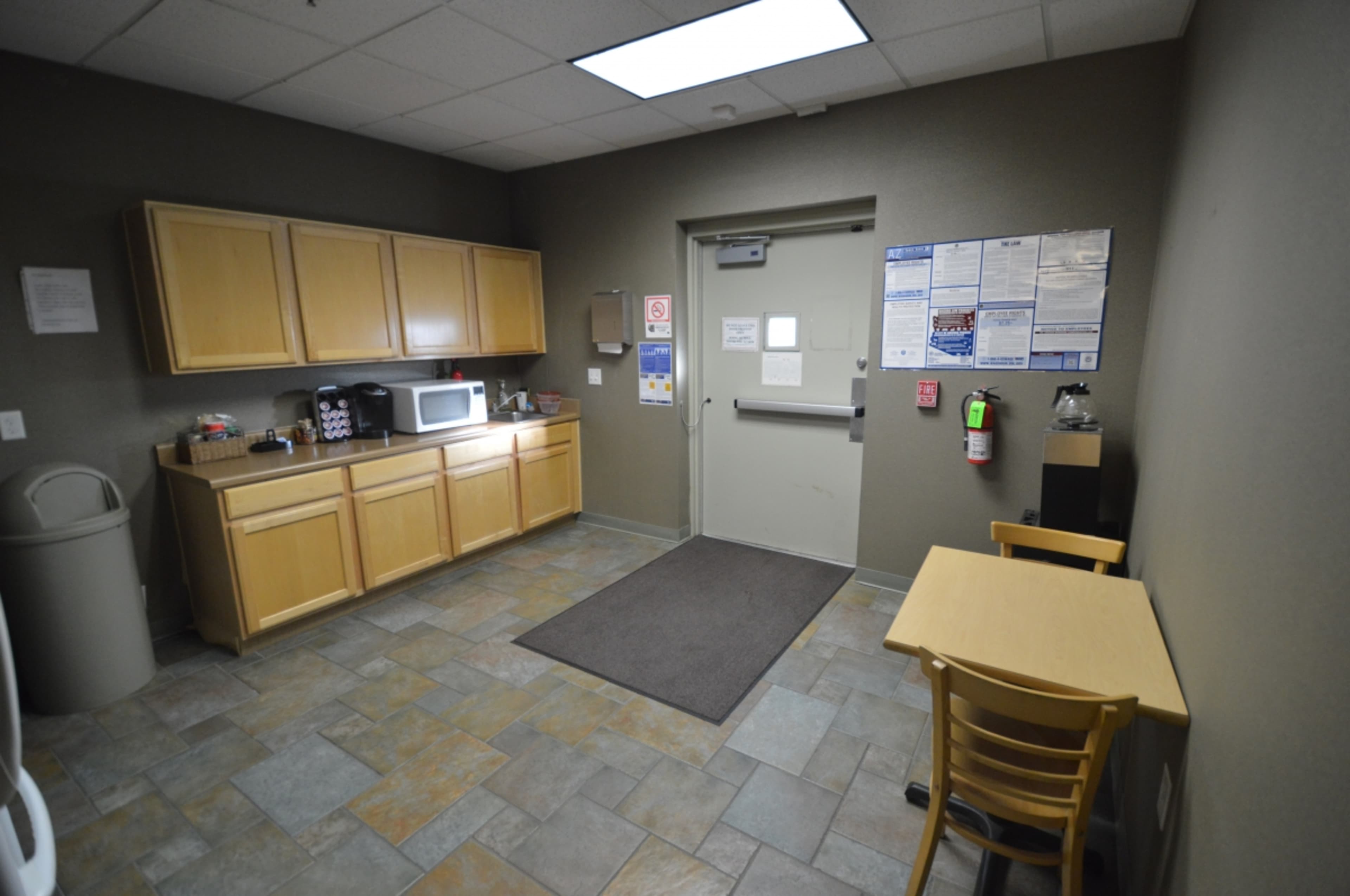 A break room with wooden cabinets, a microwave, a small table with a chair, and a door leading to the outside.