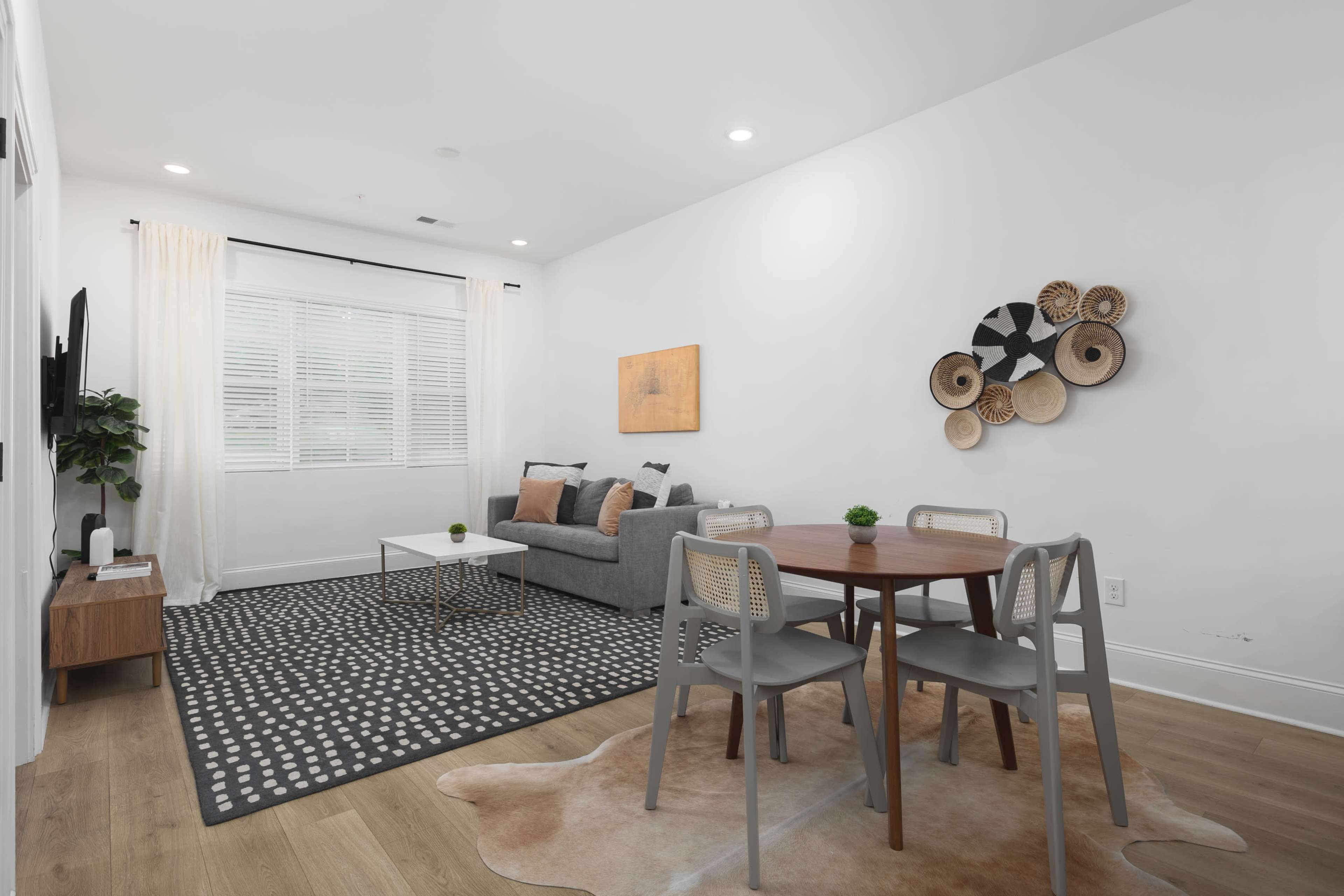 The image shows a modern living and dining area featuring a gray sofa, a wooden dining table with chairs, and decorative wall art.