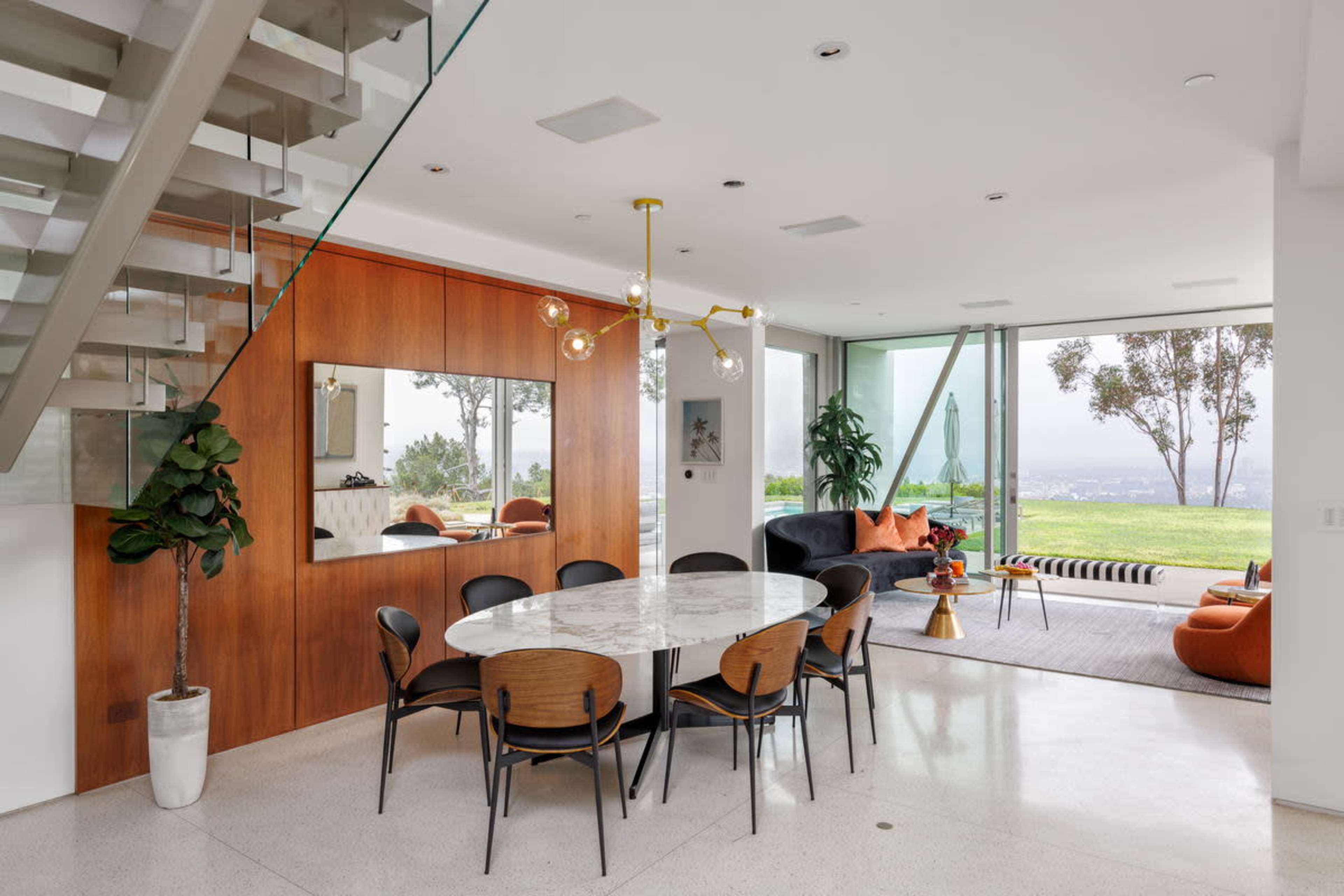 A modern dining area features a round marble table surrounded by black and wooden chairs, with large windows showcasing a view and a staircase in the background.