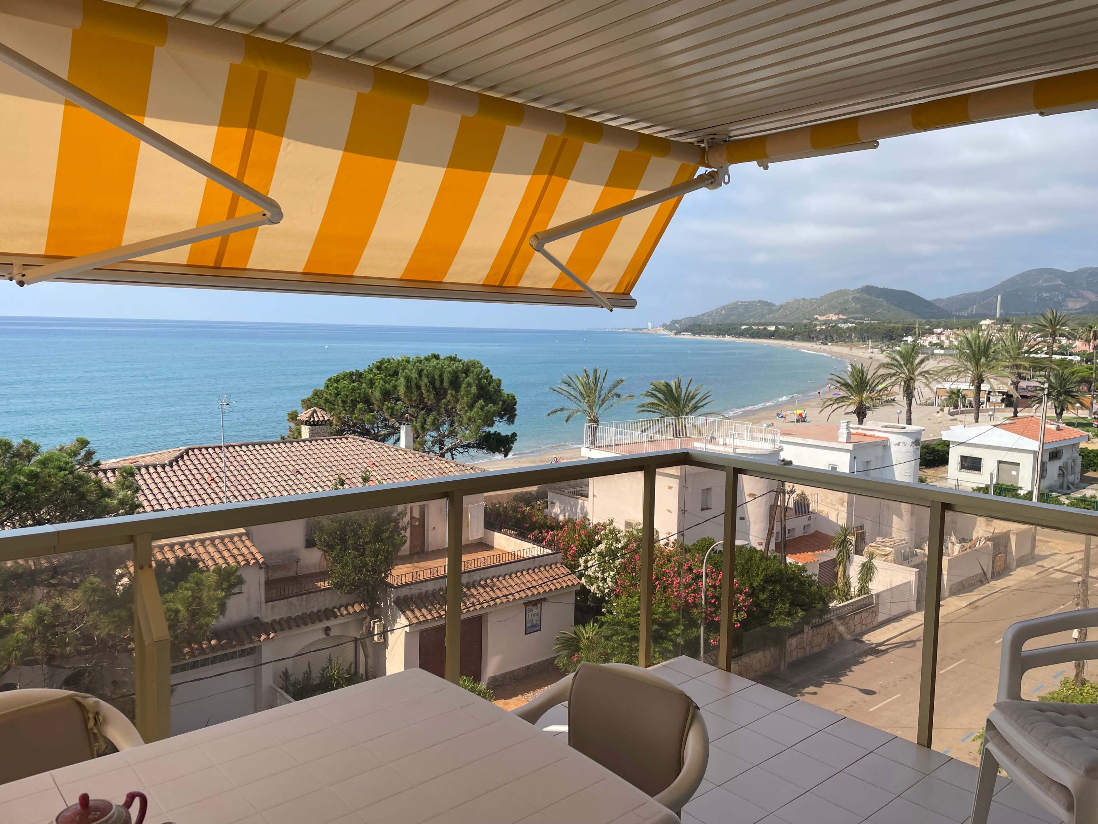 The image shows a balcony view overlooking a beach with calm waters, palm trees, and distant mountains under a partly cloudy sky.