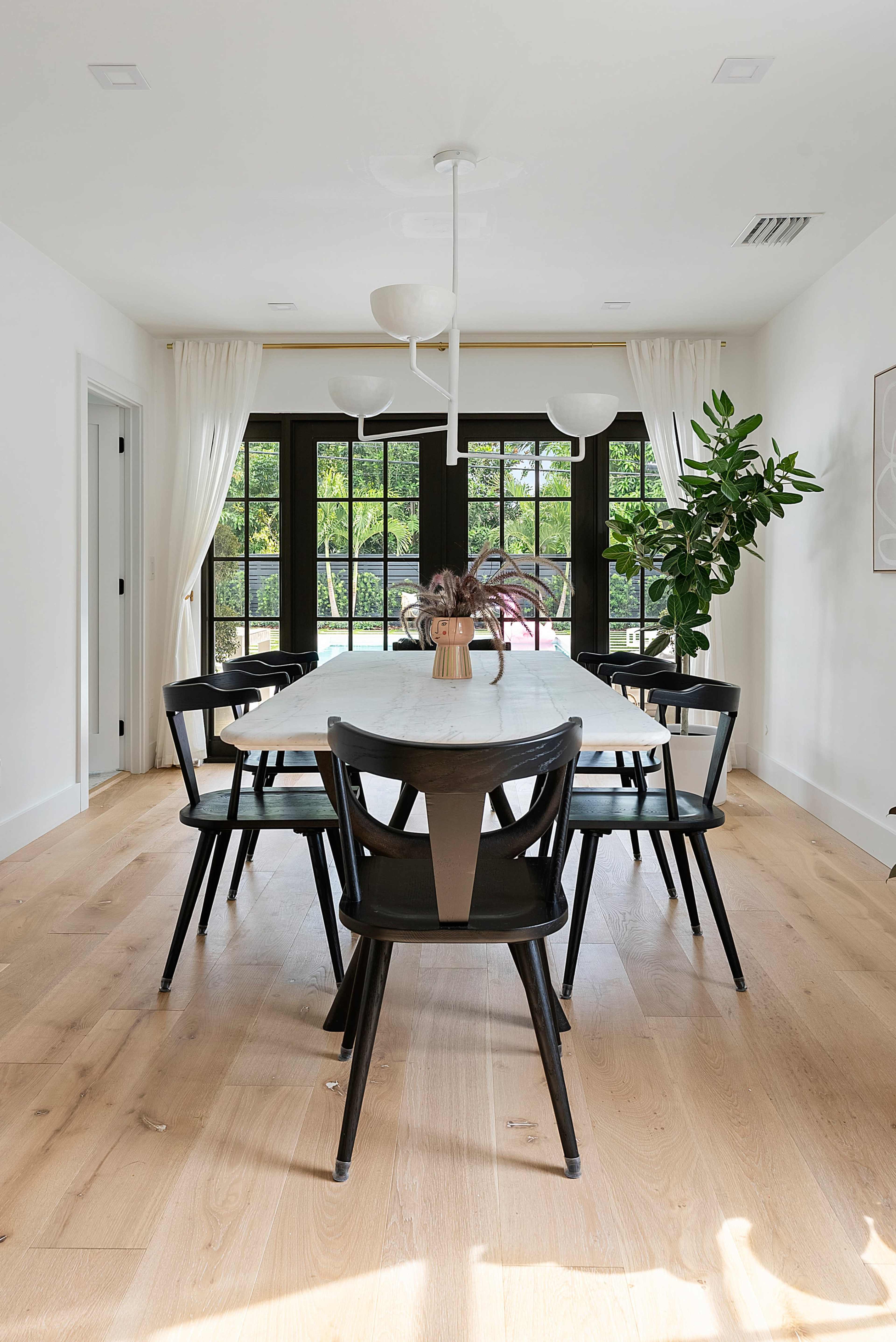 A modern dining room features a marble table surrounded by black chairs, with large windows allowing natural light and a view of greenery outside.