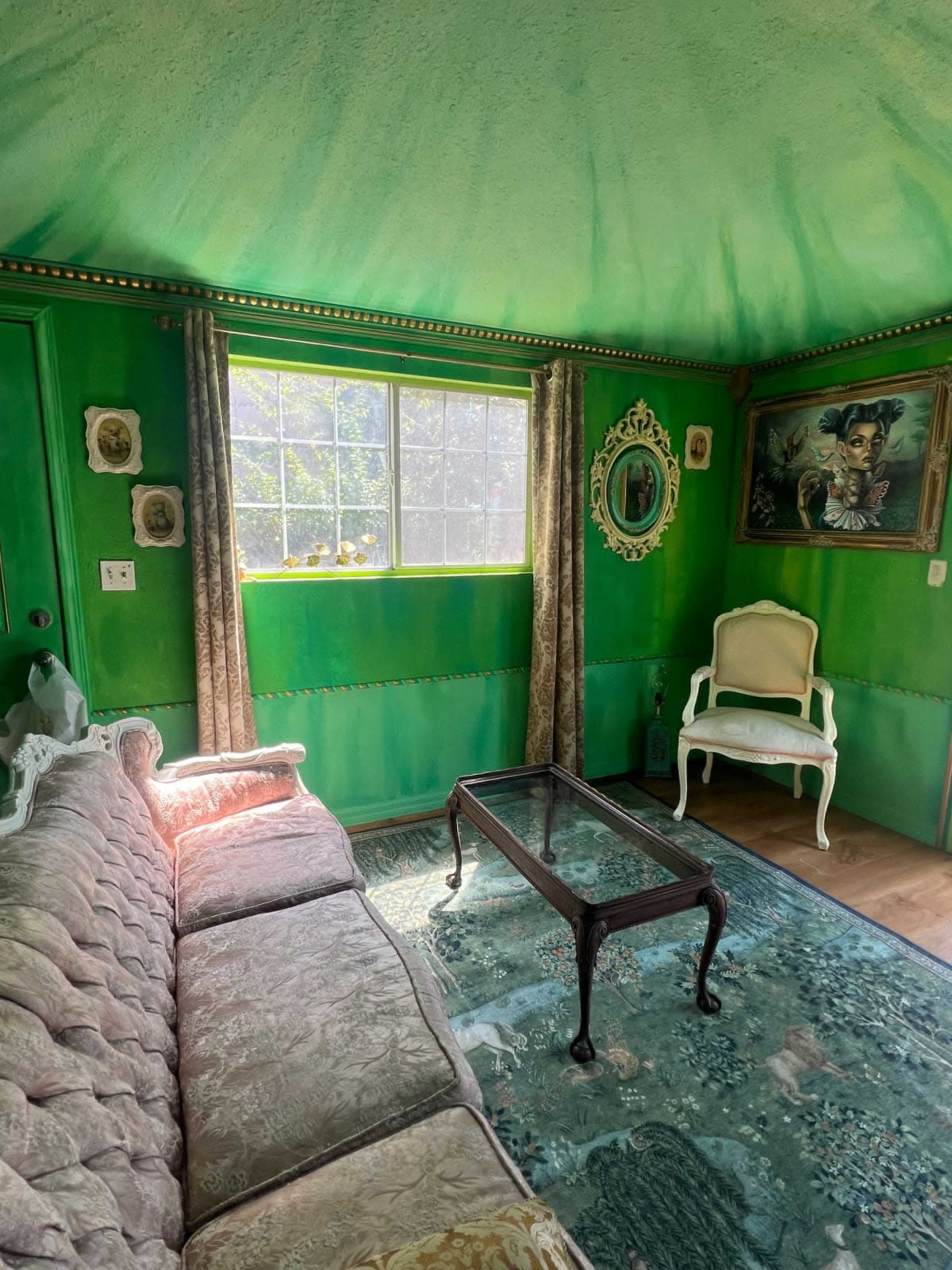 A green-walled room with a patterned sofa, a glass coffee table, and a window letting in natural light.