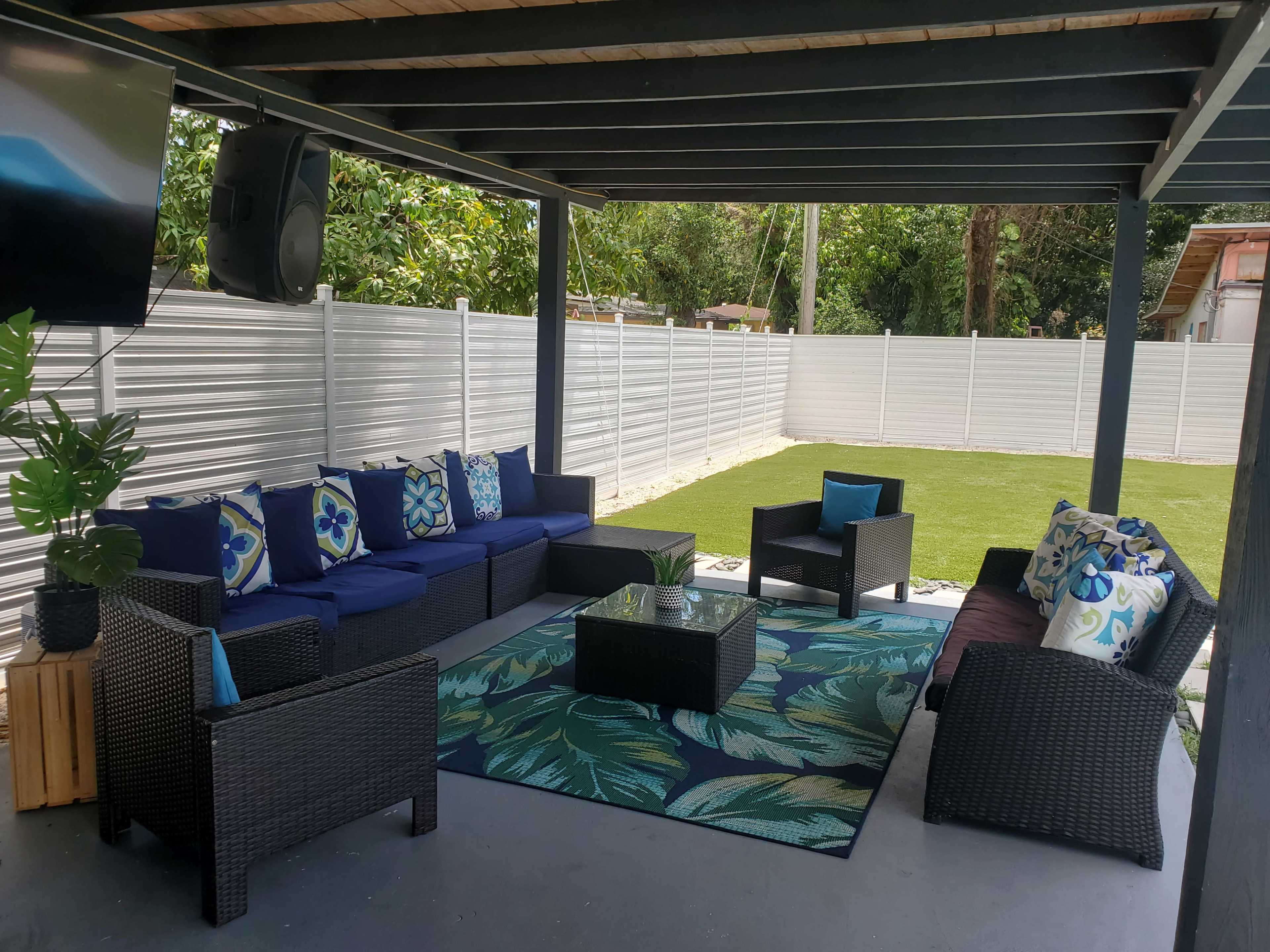The image shows a covered outdoor seating area with a large sectional couch, a glass coffee table, and a vibrant area rug, surrounded by lush greenery and a white fence.
