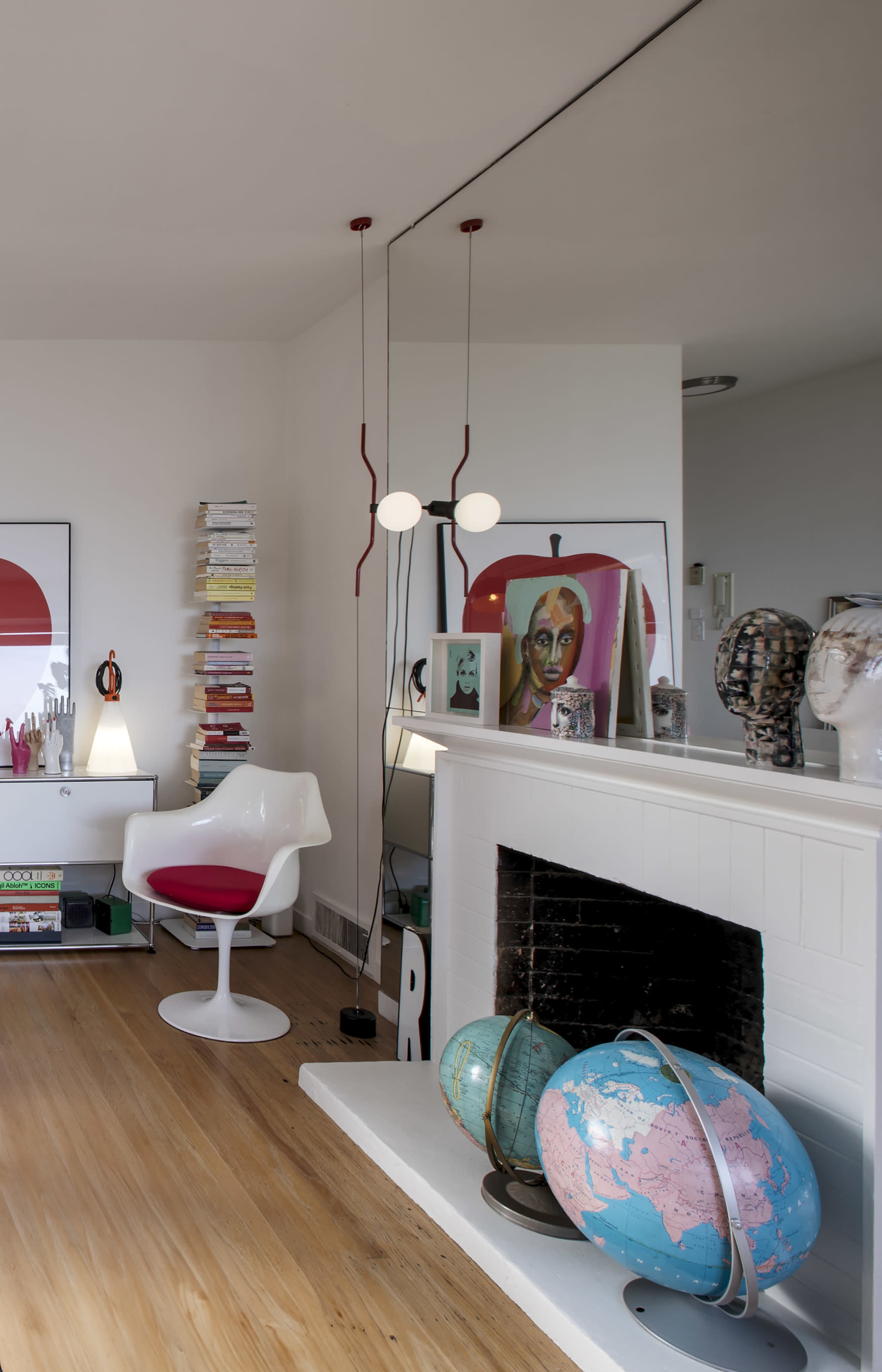 The image shows a modern living room with a white chair, a bookshelf filled with stacked books, a decorative fireplace, artwork on the walls, and a globe on a stand.