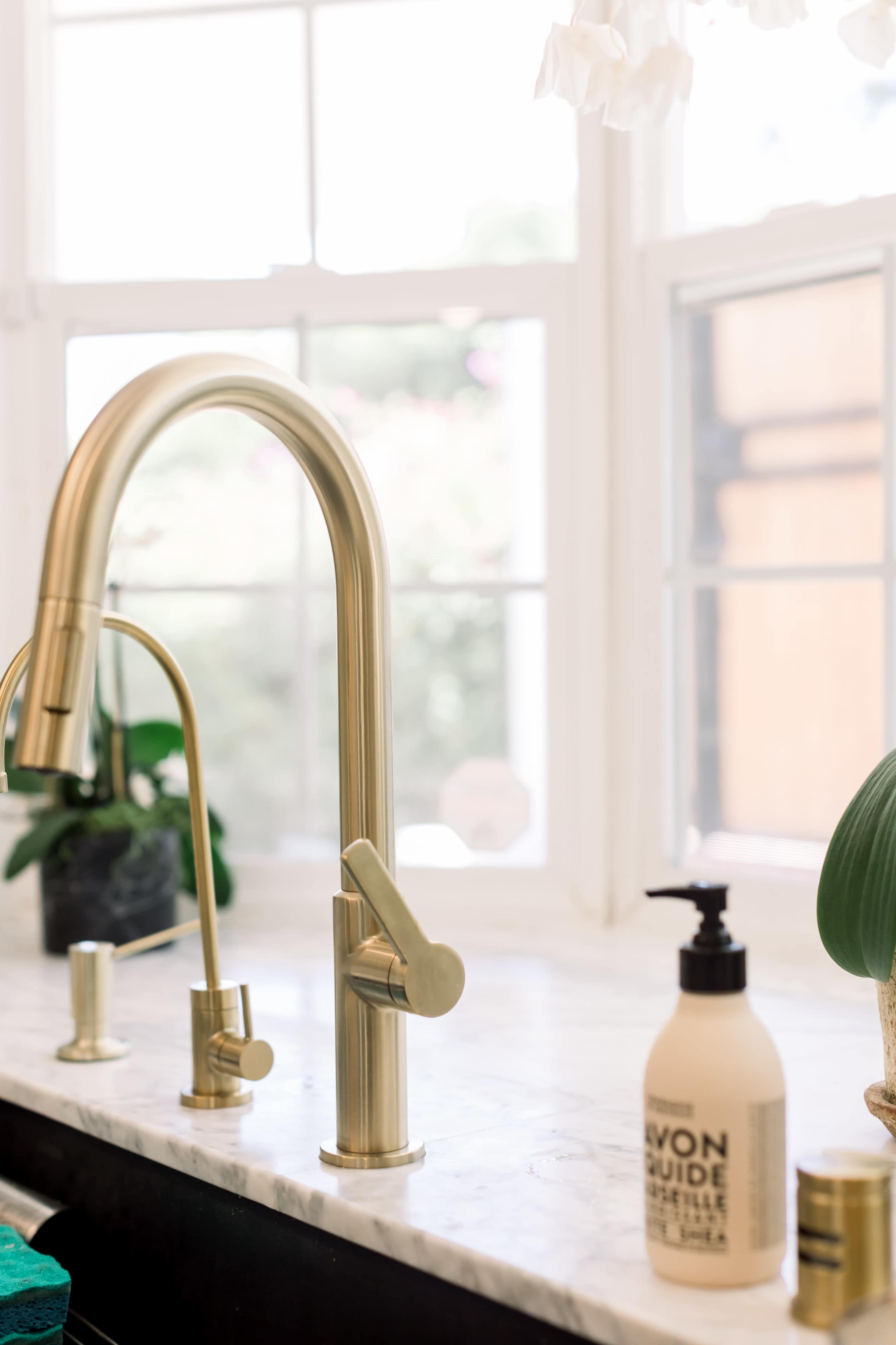 A brass kitchen faucet and soap dispenser on a marble countertop near a window.
