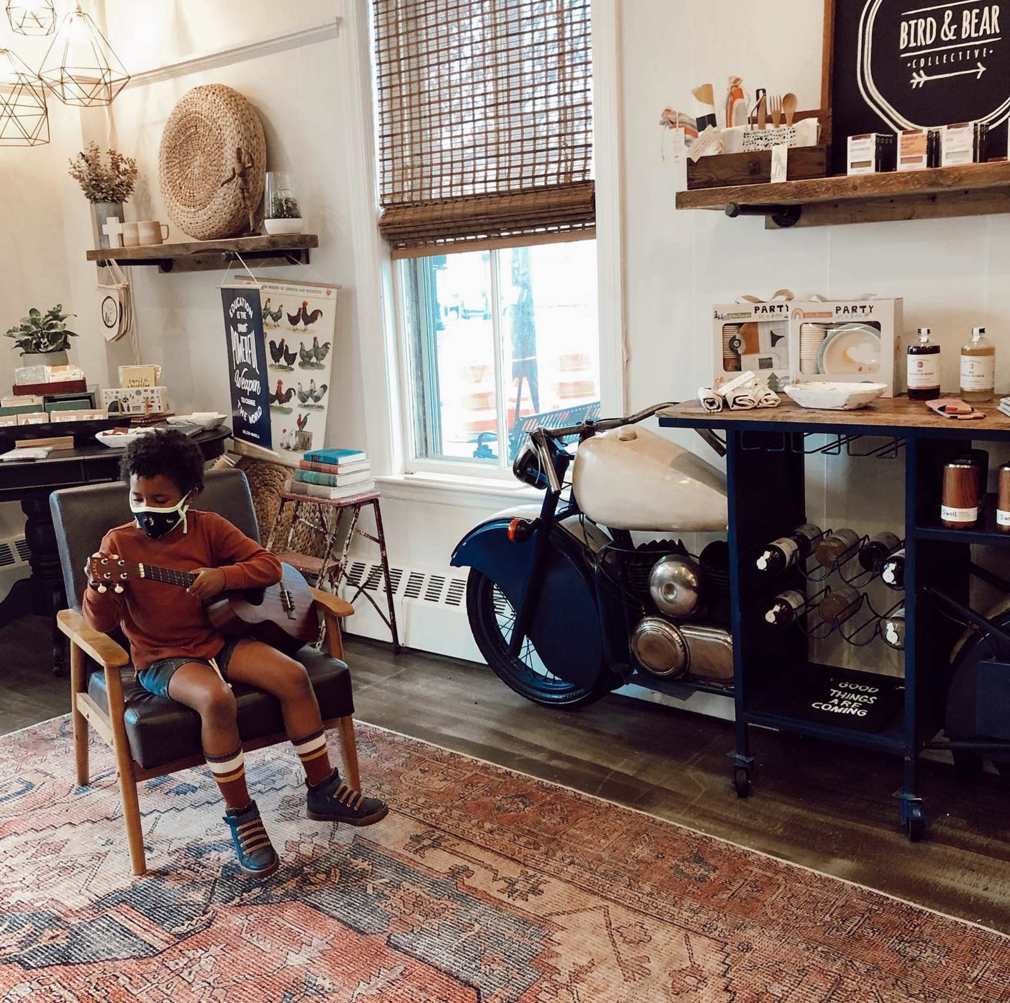A child sits on a chair playing a ukulele in a cozy shop filled with home decor and a vintage motorcycle.