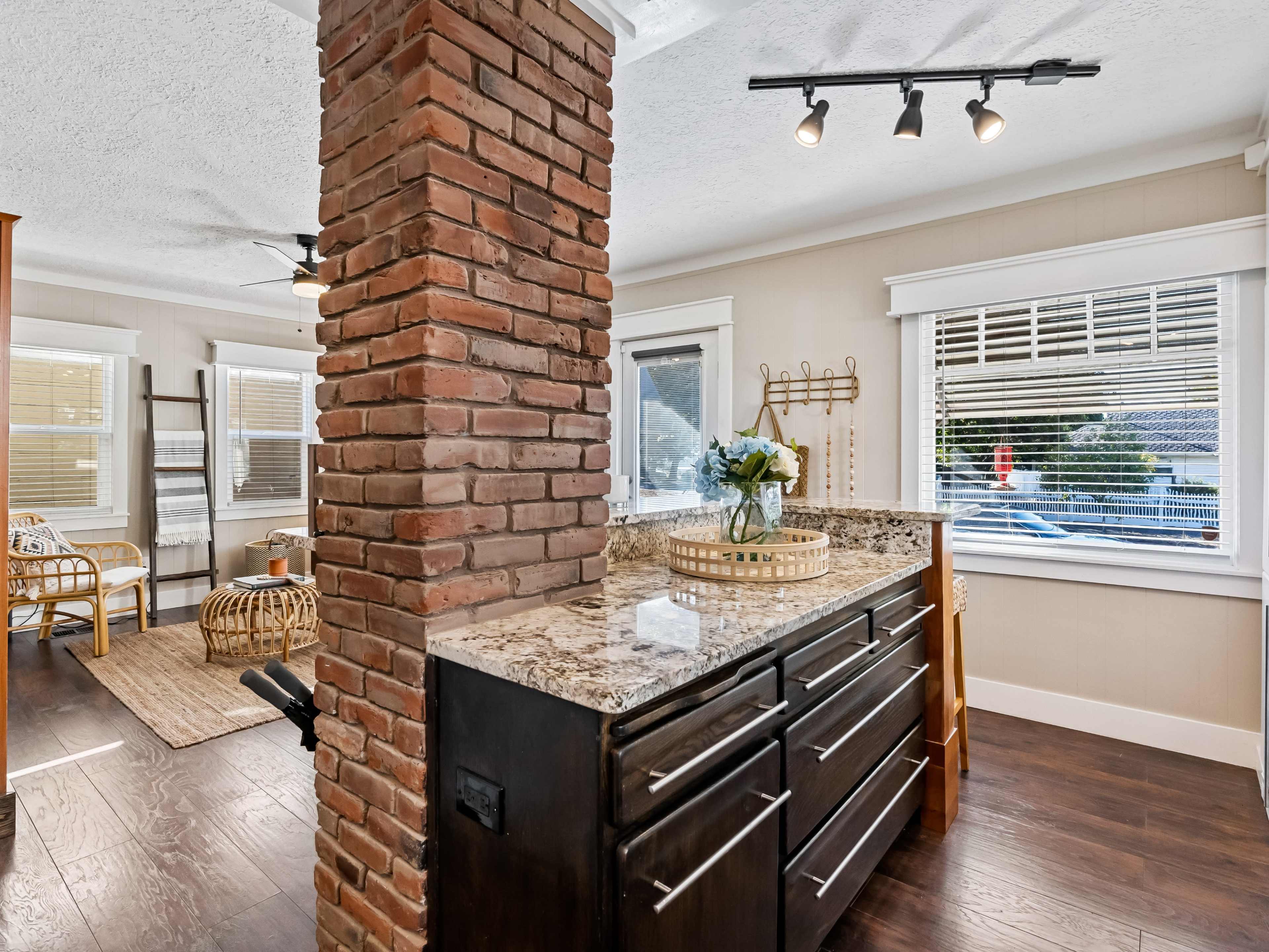 A modern kitchen with a granite countertop and a brick column separating it from a cozy seating area featuring wicker furniture and large windows.