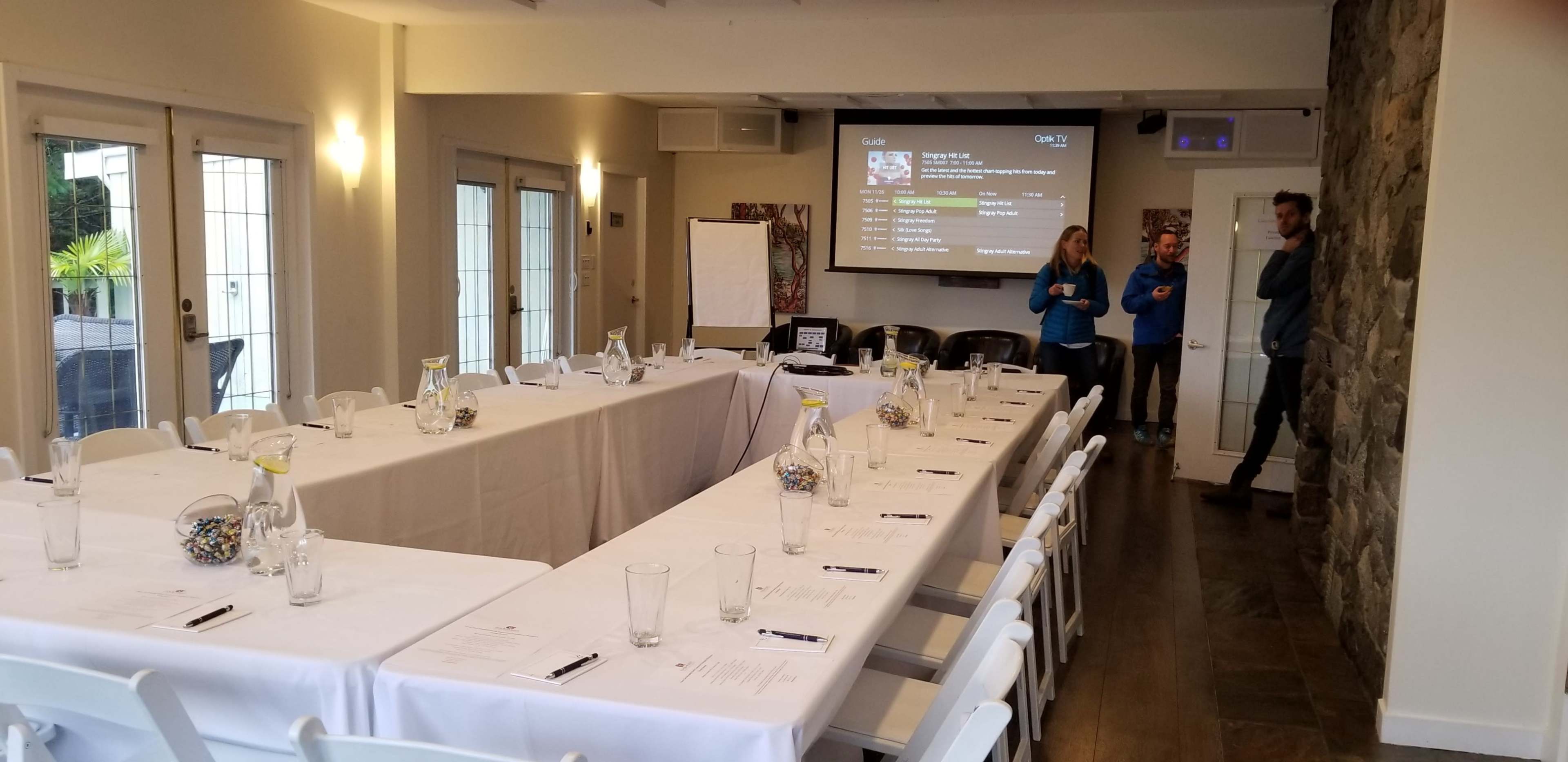 The image shows a conference room with a long table set for a meeting, featuring notepads and glasses, while two individuals stand near a presentation screen displaying information.