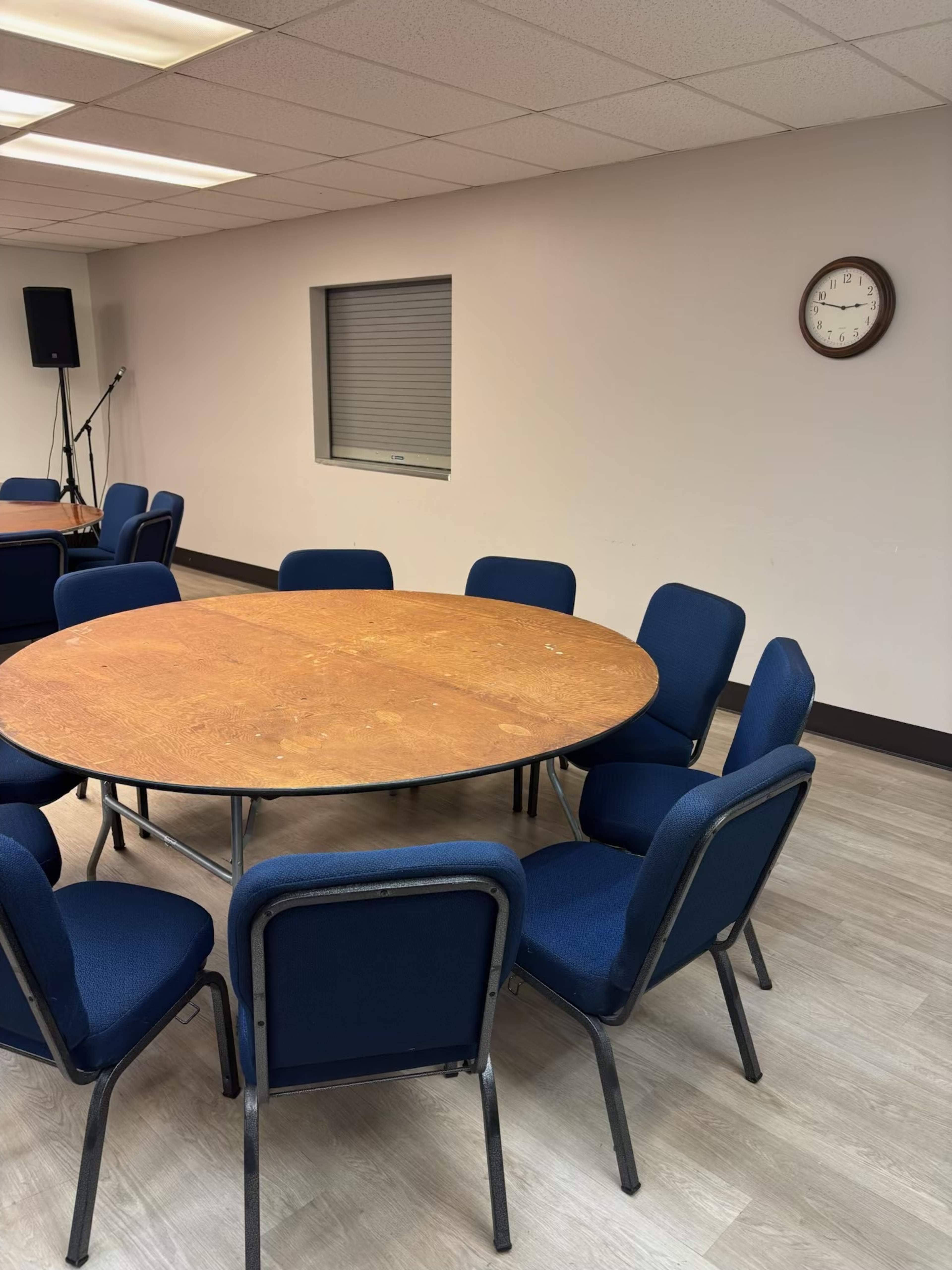 A round wooden table surrounded by blue chairs is positioned in a room with a clock on the wall and a window covered by blinds.