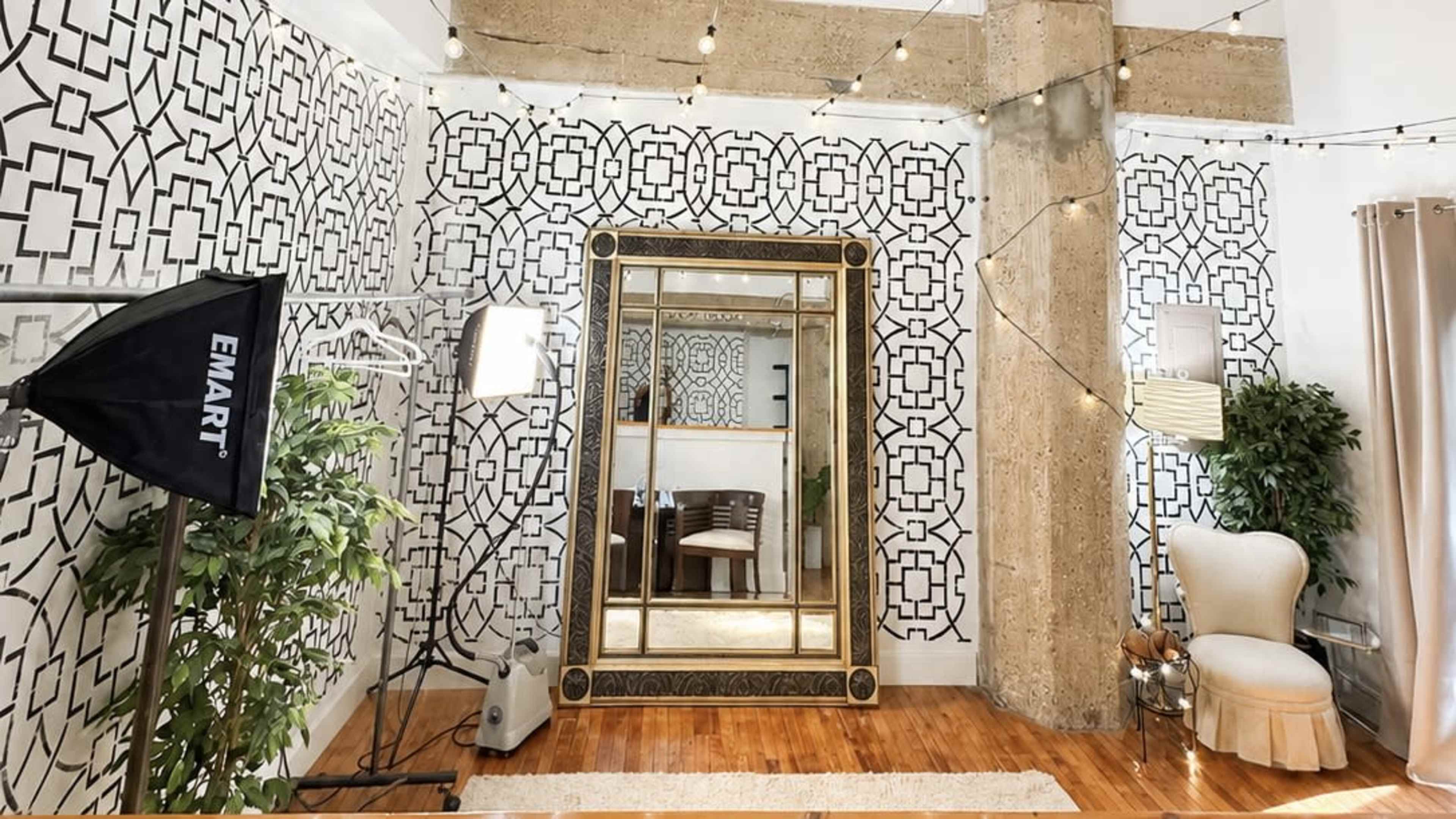 The image shows a spacious room with patterned wallpaper, a large ornate mirror, and a light-colored chair, complemented by soft lighting and decorative plants.