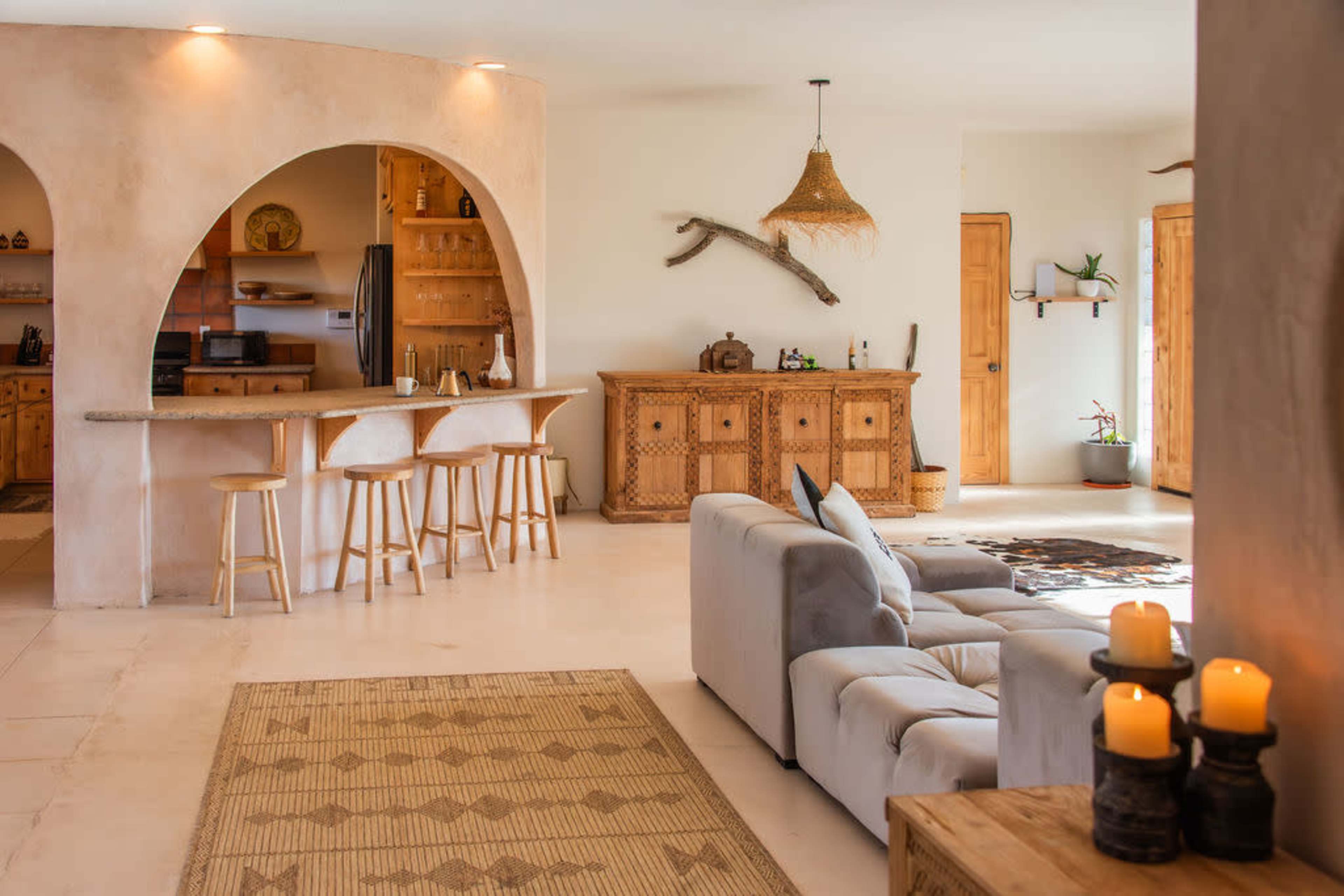The image shows a spacious living area featuring a kitchen bar with stools, a sofa, and a decorative wooden cabinet, all within a light-colored room.