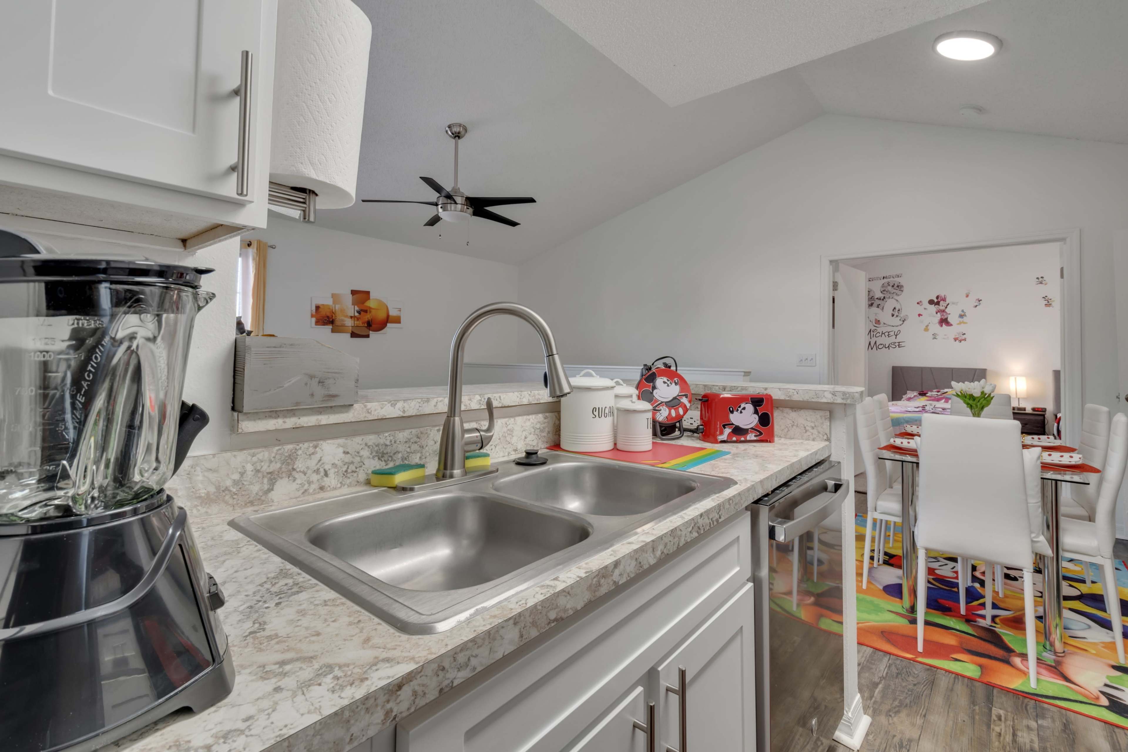 A modern kitchen features a double sink, a blender on the counter, and a view towards a dining area with colorful decor.