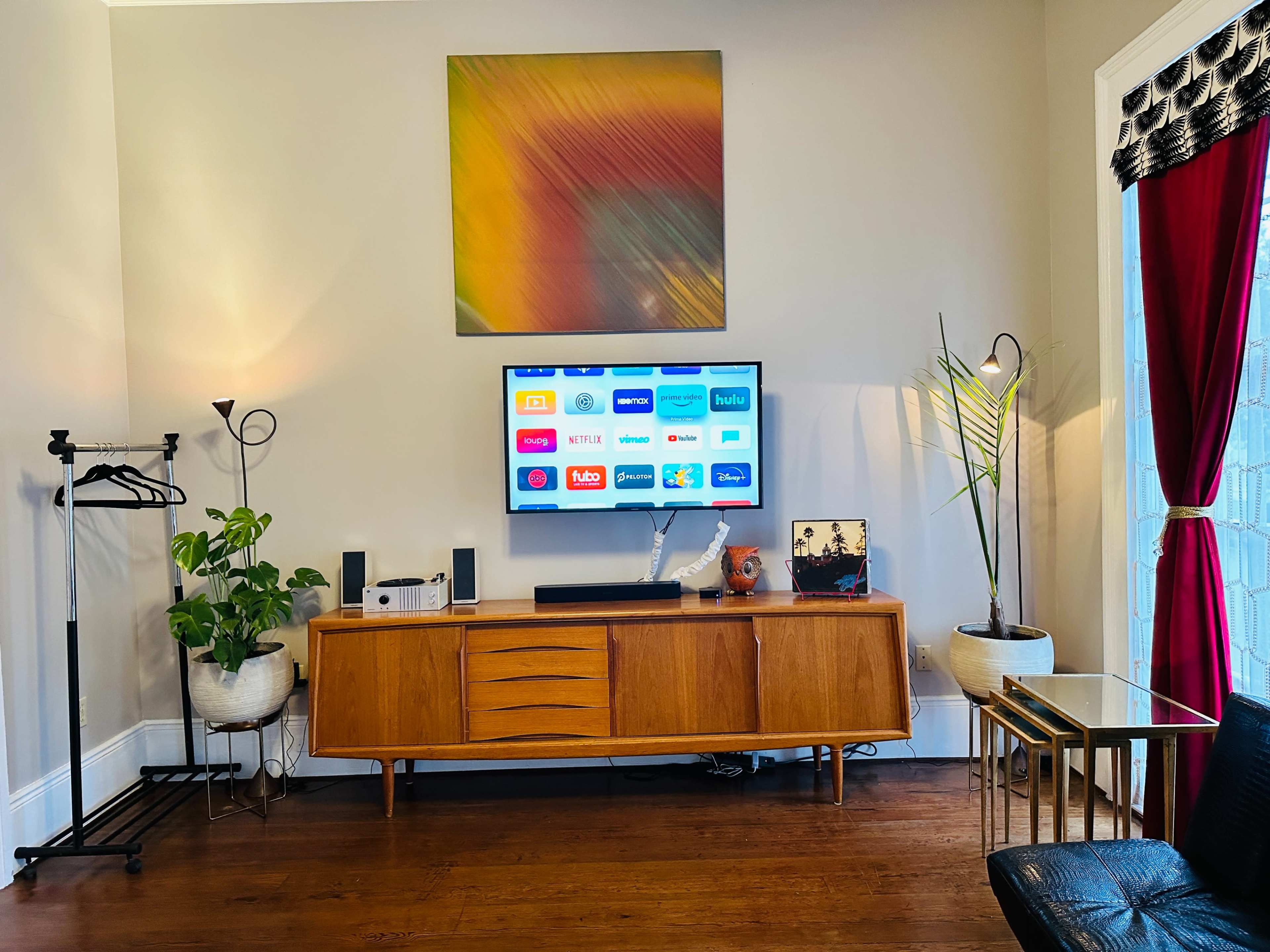 A living room features a wooden media console with a TV displaying various streaming app icons, surrounded by indoor plants and decorative items, with a coat rack and floor lamp nearby.