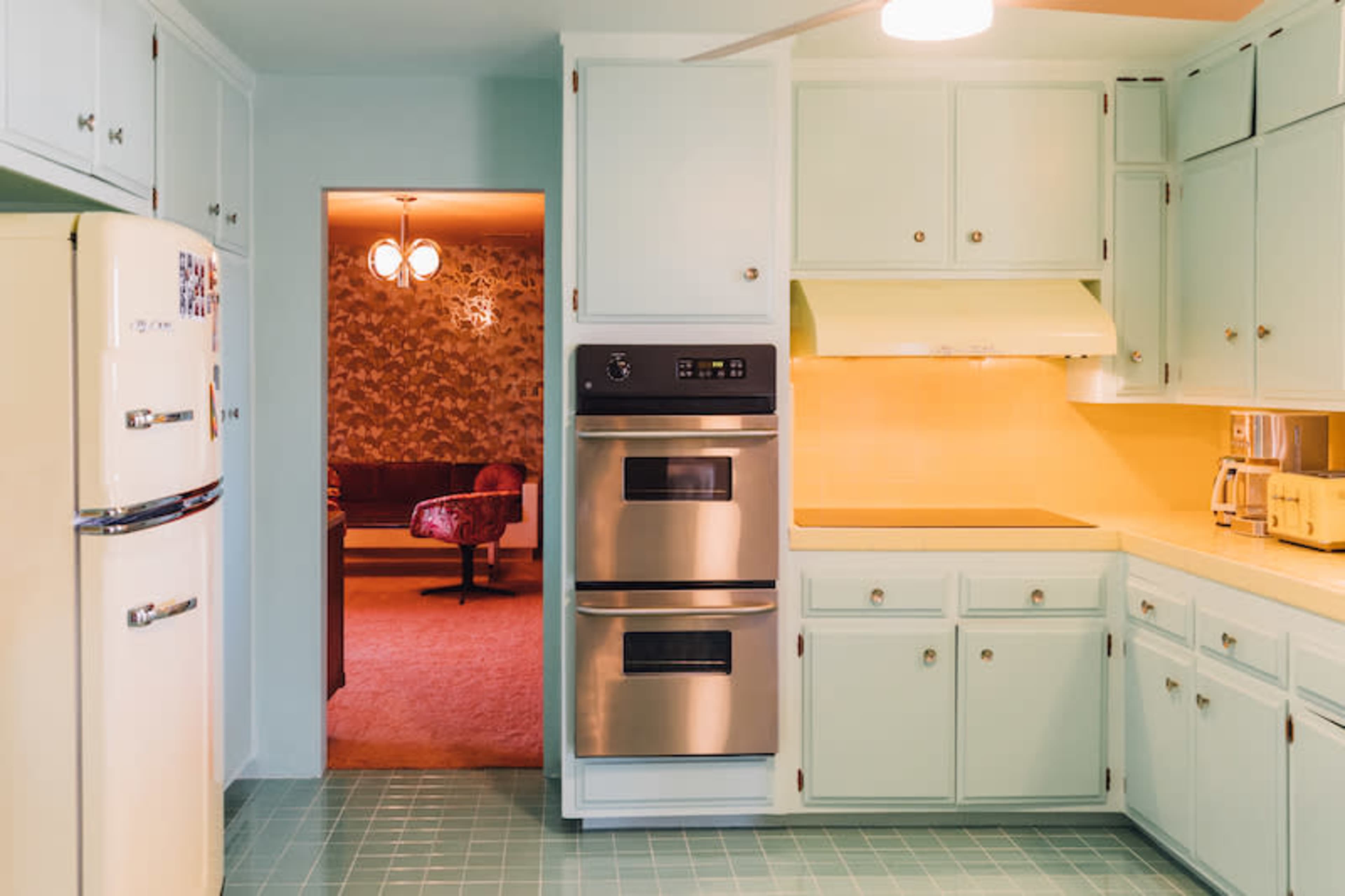 The kitchen features light blue cabinets, stainless steel appliances, and an open doorway leading to a living area with patterned wallpaper.